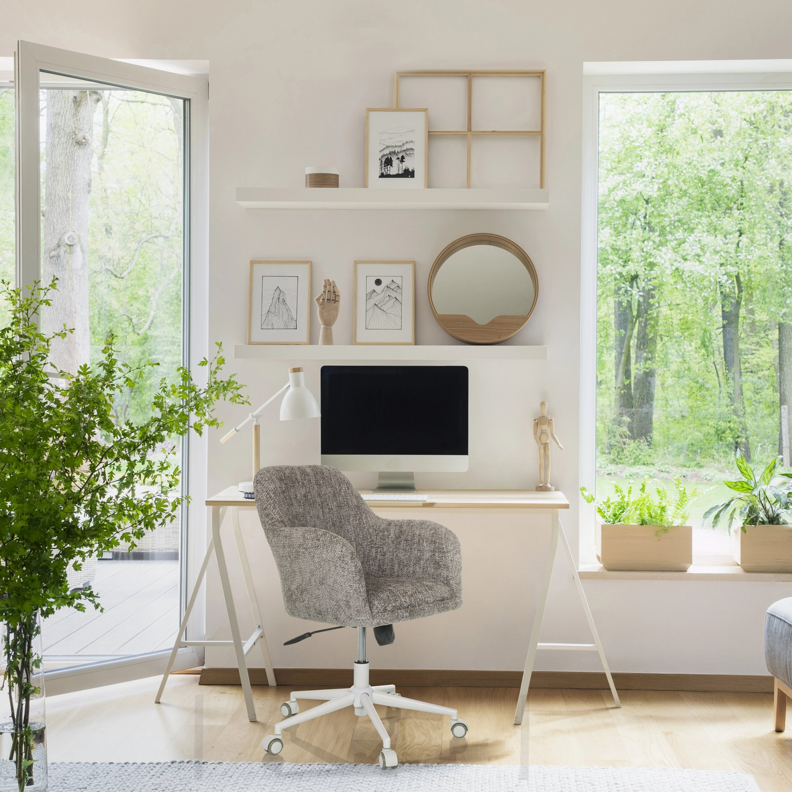Ein heller Arbeitsbereich mit einem Schreibtisch, Computer, grauem Stuhl und Regalen mit Dekoration vor Fenstern.
