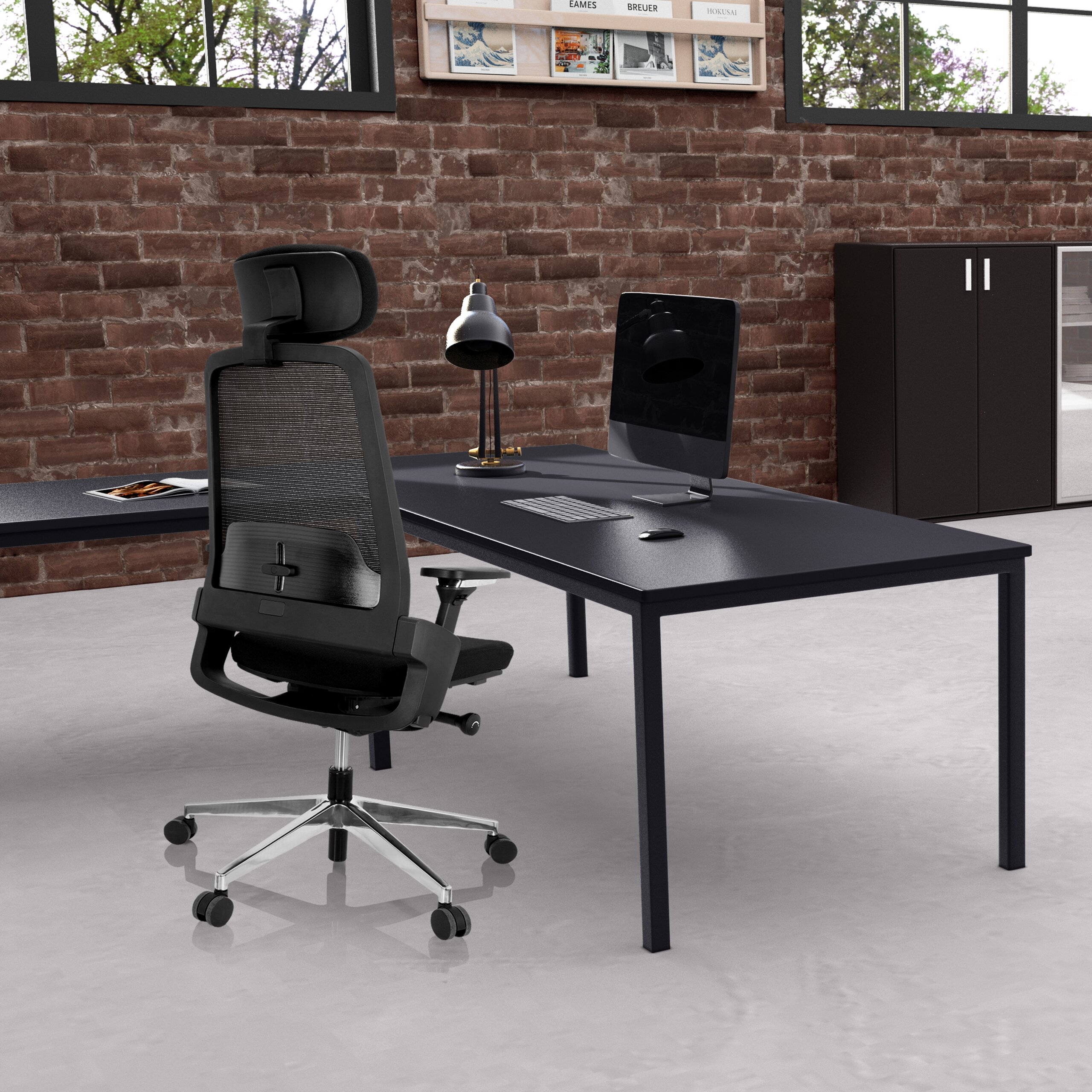 Ein schwarzer Netz-Bürostuhl steht vor einem dunklen Schreibtisch mit Computer und Lampe vor einer Ziegelwand.