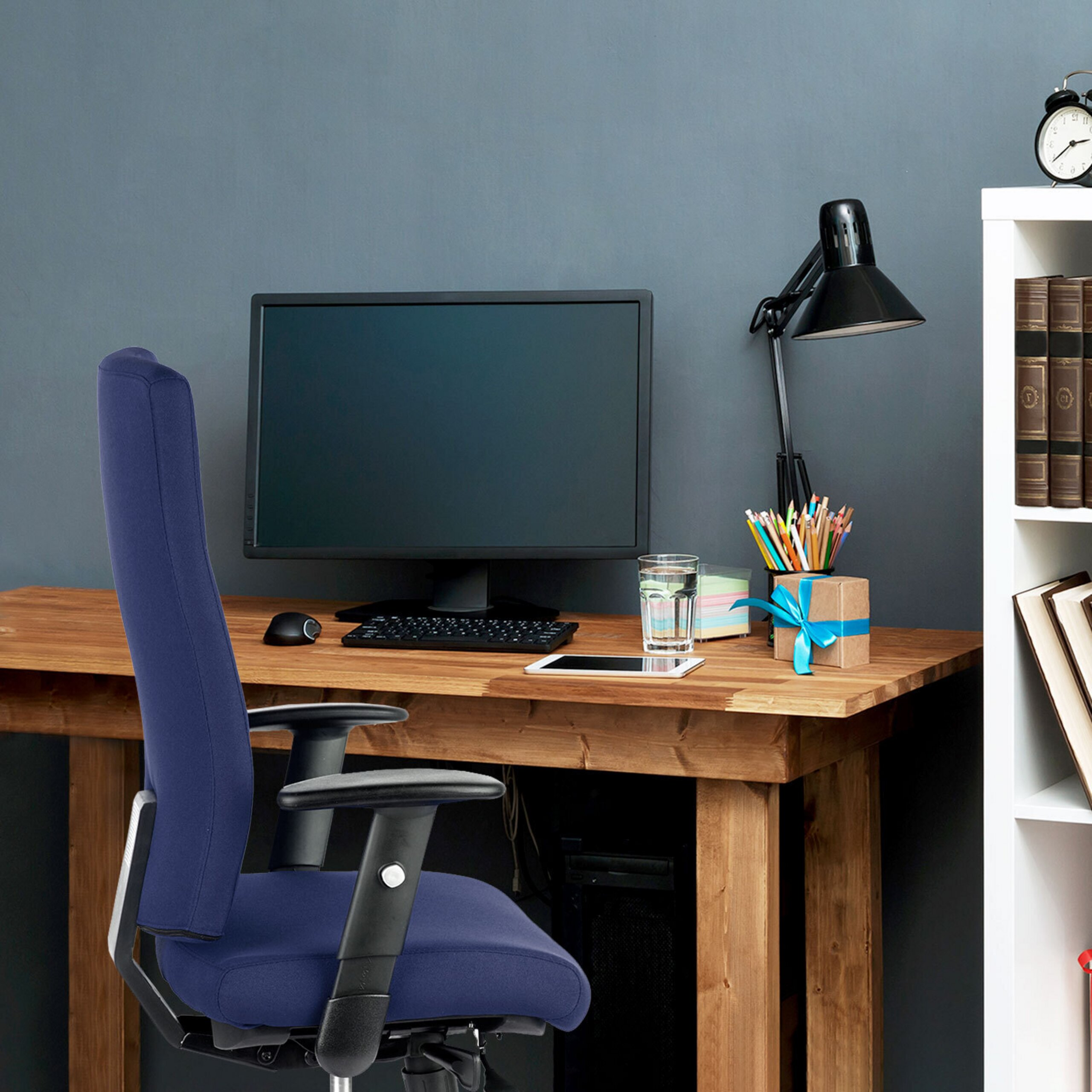 Ein Holztisch mit Monitor, Tastatur, Maus, Lampe und blauem Bürostuhl vor einer grauen Wand.