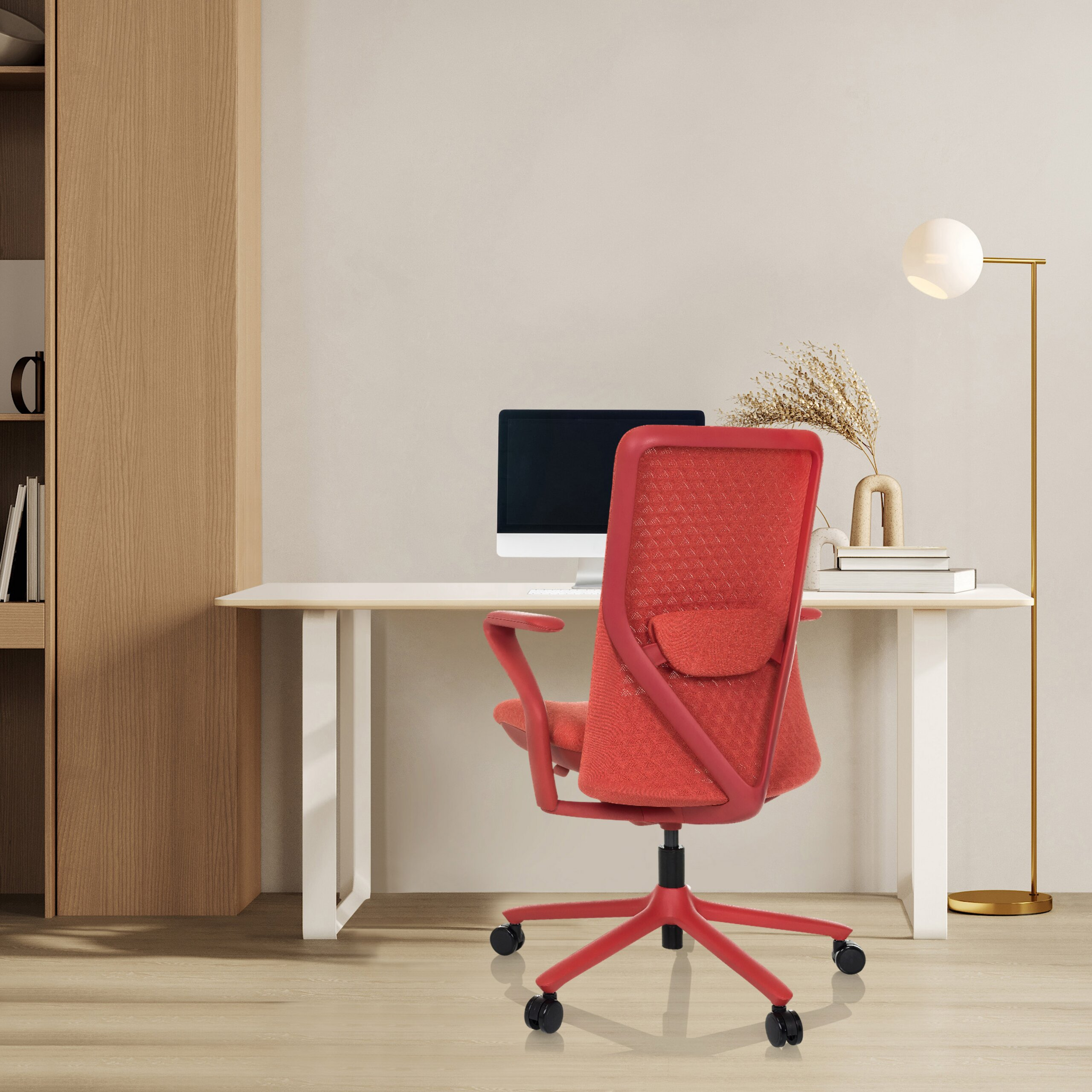 Ein roter Buerostuhl steht vor einem Schreibtisch mit Computer, Lampe und Buechern vor einer hellen Wand.
