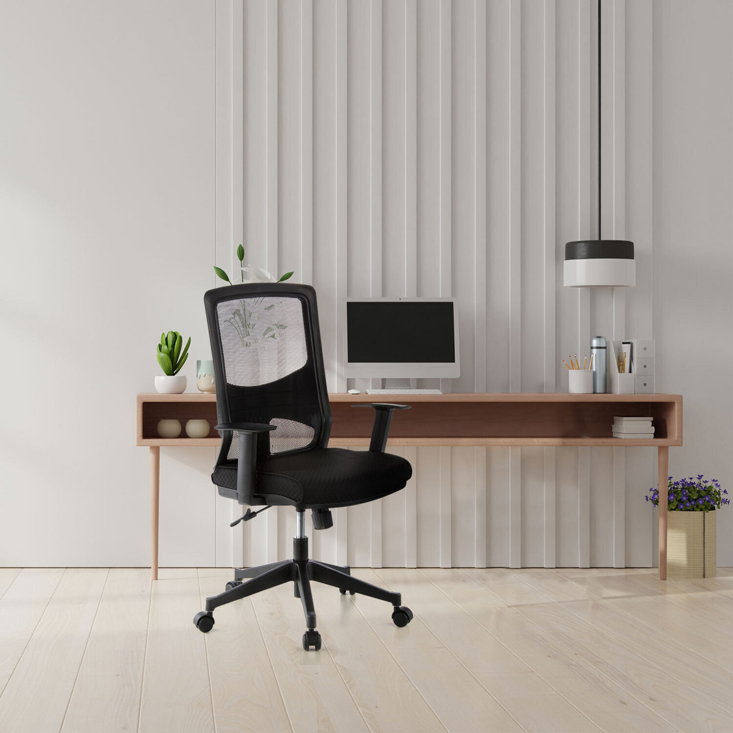 Ein schwarzer Bürostuhl vor einem Holztisch mit Computer und Pflanzen an einer weißen Paneelwand.