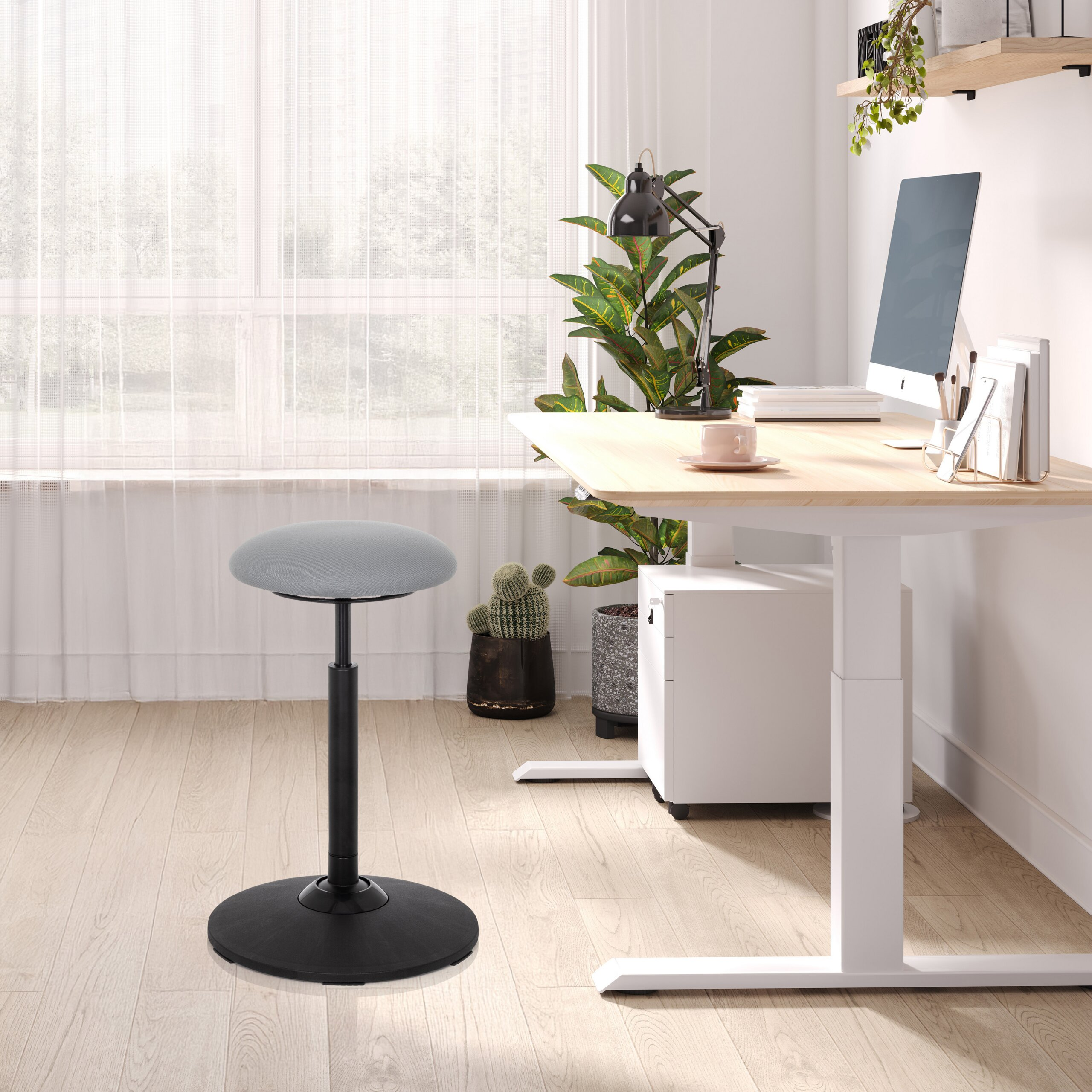 Ein grauer Stehhocker mit schwarzem Fuß steht vor einem Holztisch mit Computer, Pflanzen und Fenster.