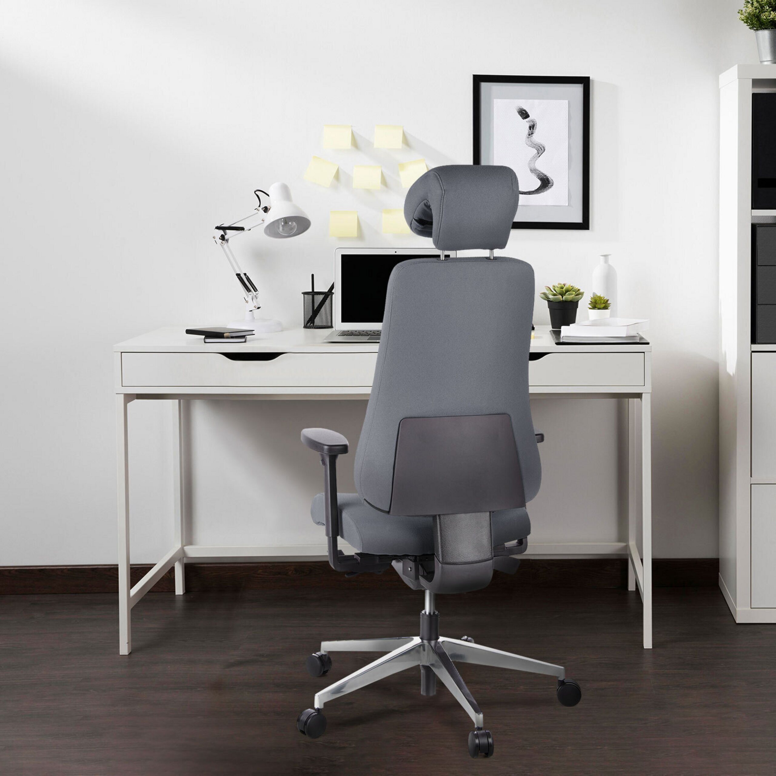 Ein ergonomischer grauer Stuhl steht vor einem weißen Schreibtisch mit Laptop, Lampe und Notizen an der Wand.