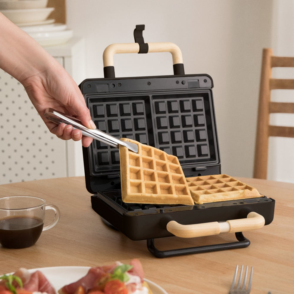 Una mano usa pinzas para levantar un gofre fresco de una gofrera moderna en una mesa de madera.