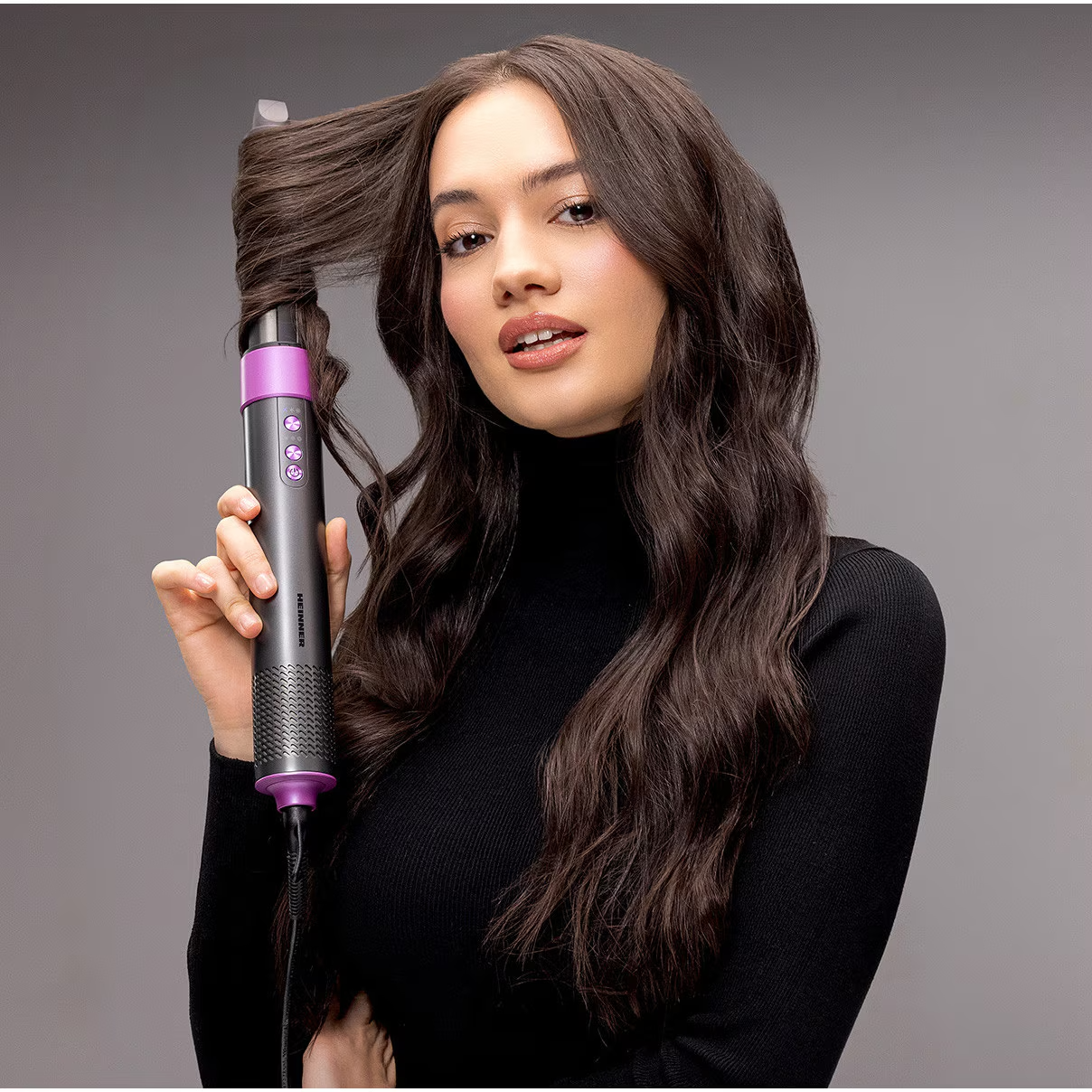 Une femme aux cheveux foncés se coiffe avec un fer à friser violet et noir sur fond gris.