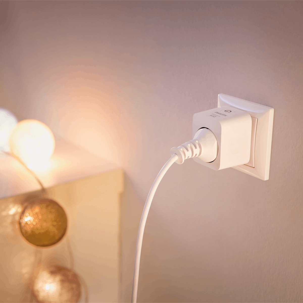 Un enchufe inteligente blanco en la pared con un cable conectado; luces de colores cálidos cuelgan a la izquierda.