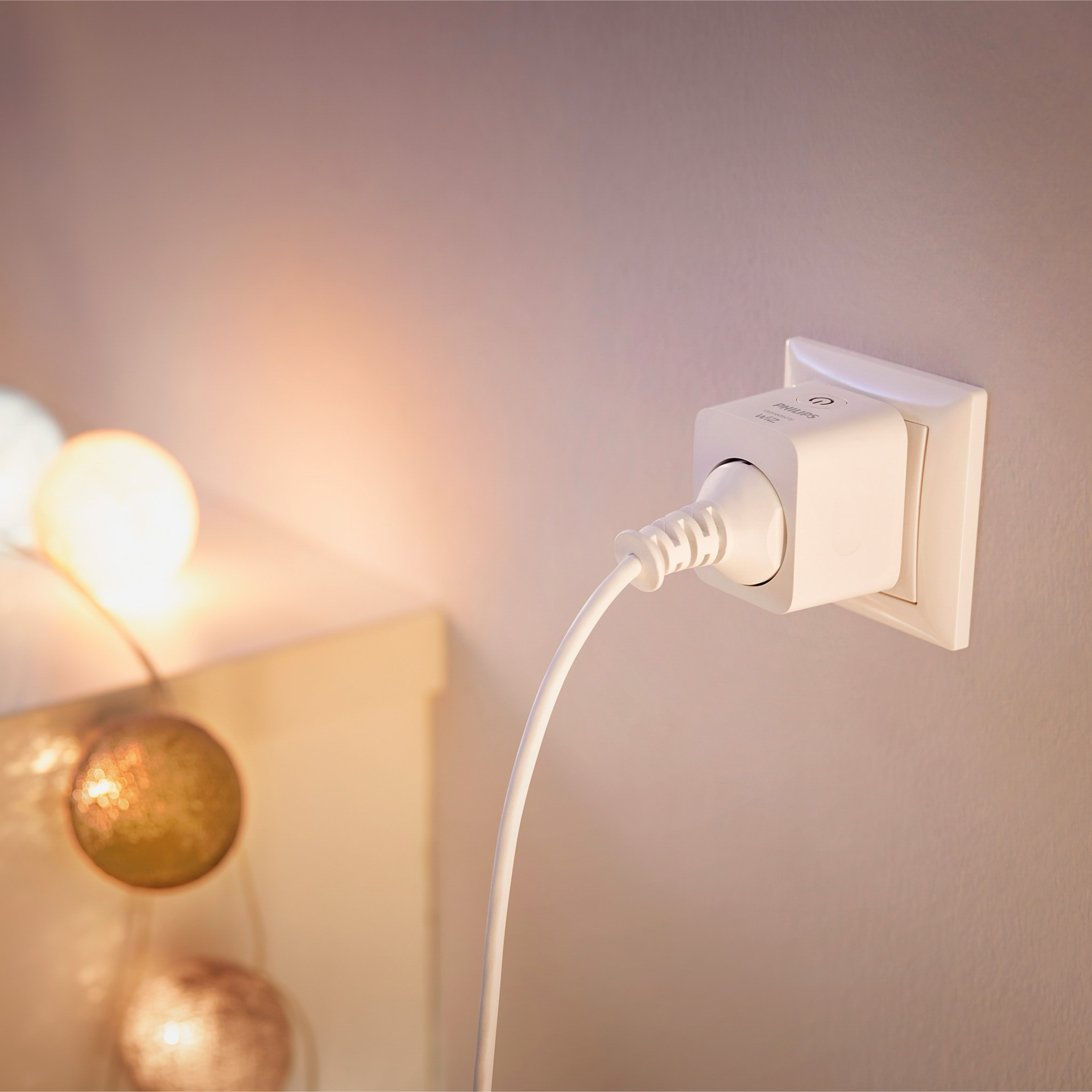 Un enchufe inteligente blanco está conectado a la pared con un cable. Una cadena de luces brilla suavemente en el fondo.