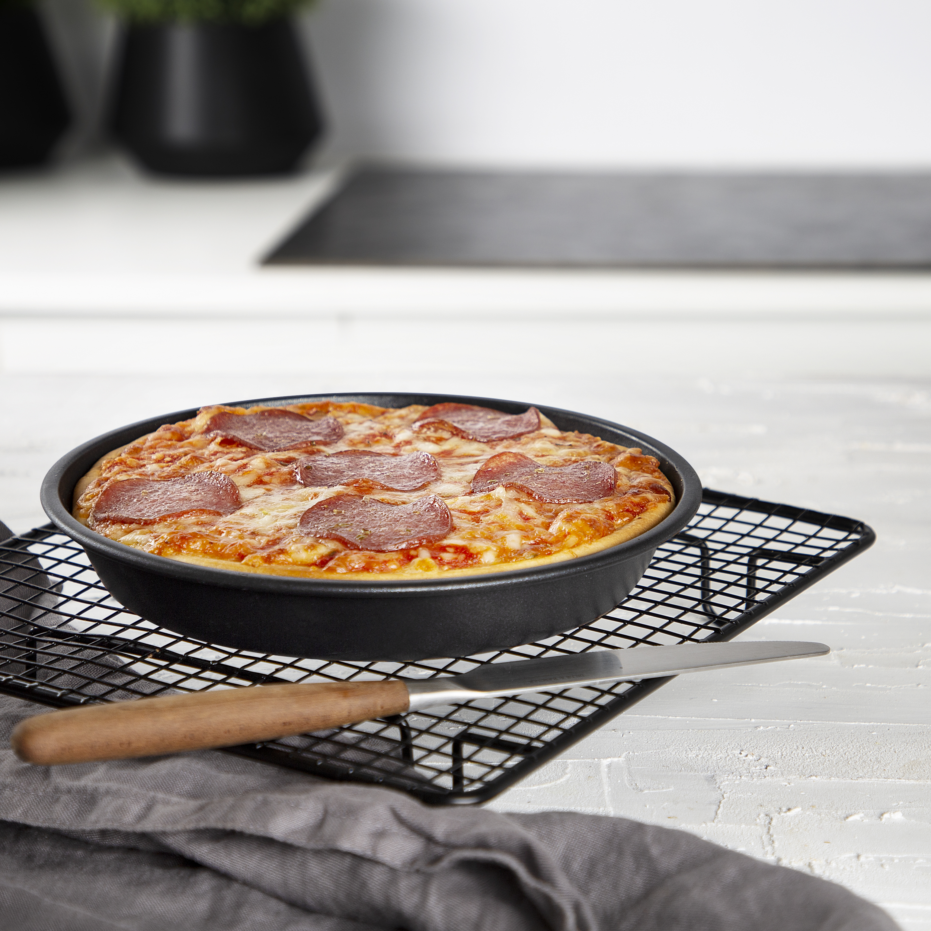 Una pizza al salame appena sfornata in una padella nera rotonda su una griglia, con un coltello e un panno grigio.