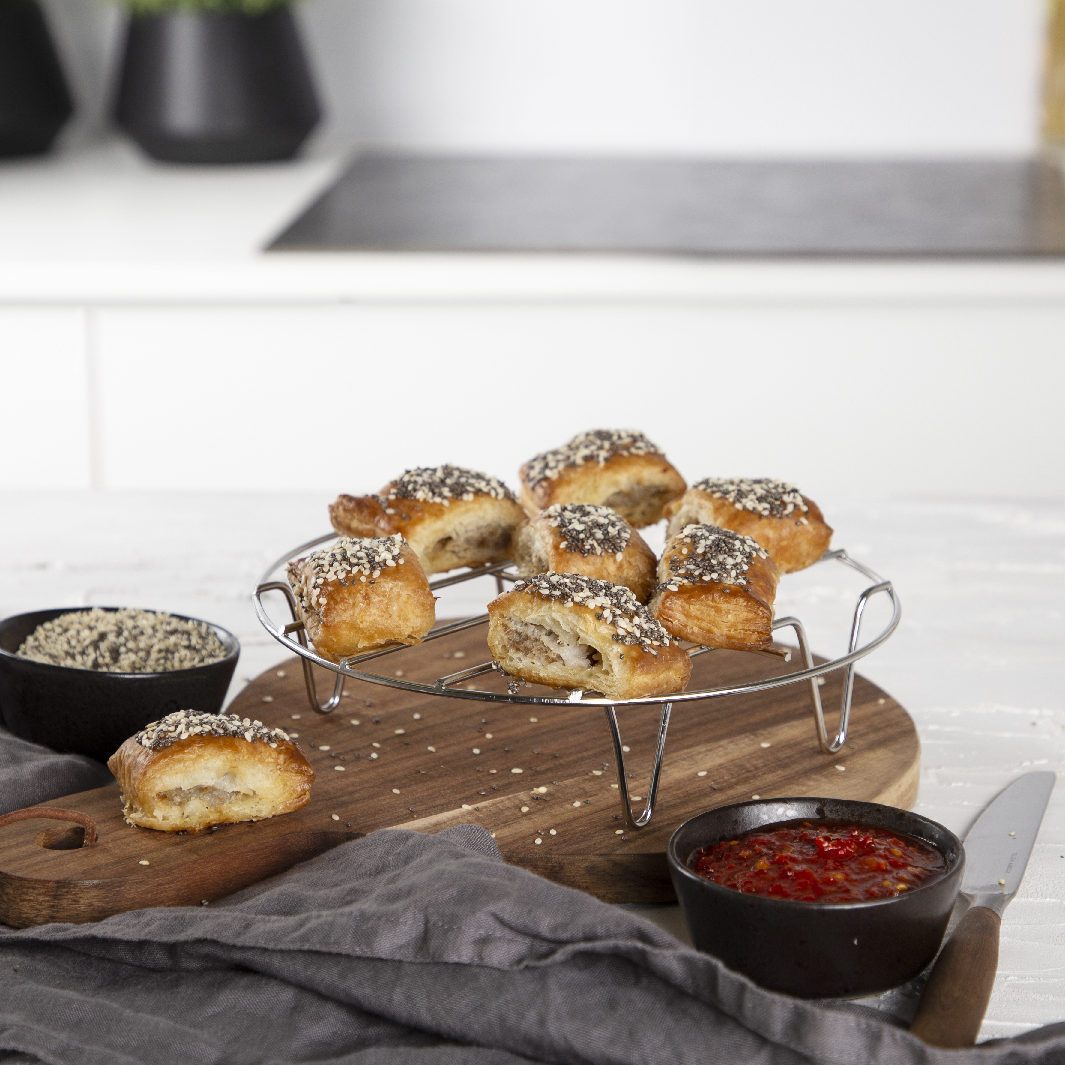 Rotolini di pasta dorata con semi su griglia e tagliere di legno, con ciotola di salsa rossa e semi.