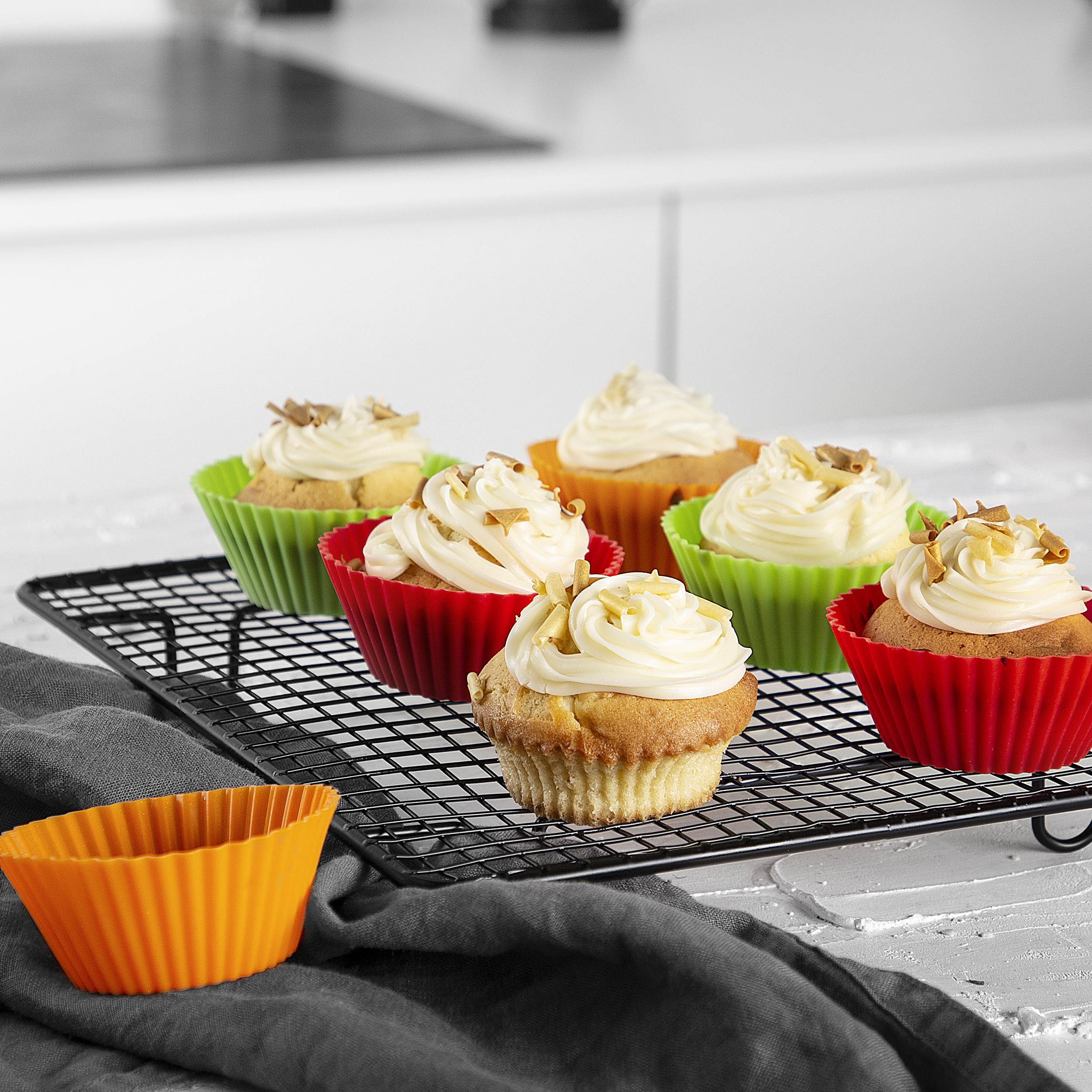 Sei cupcake con glassa bianca in pirottini colorati su una gratella in una cucina.
