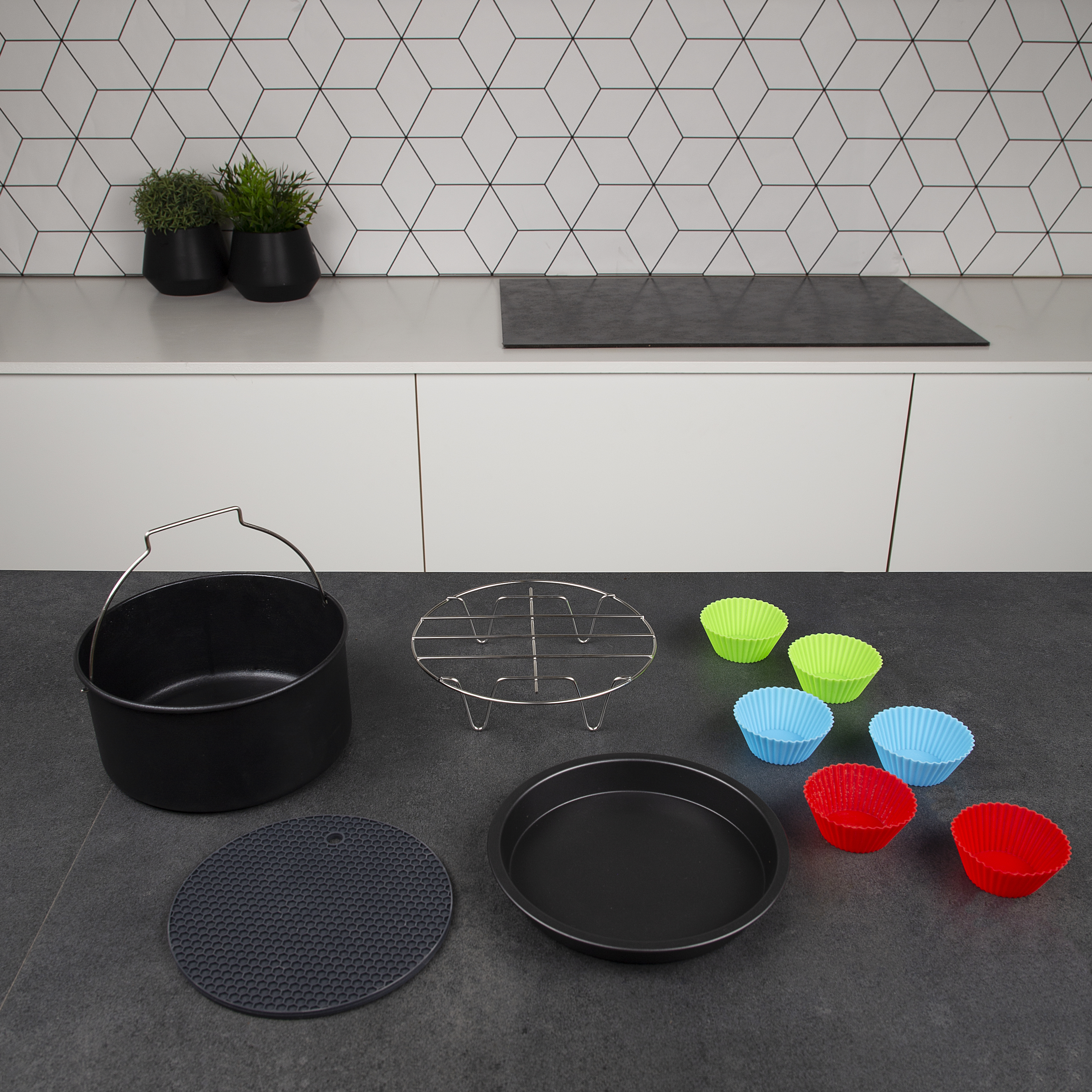 Piano cucina con teglia nera, griglia, tappetino e stampi in silicone colorati su parete bianca.