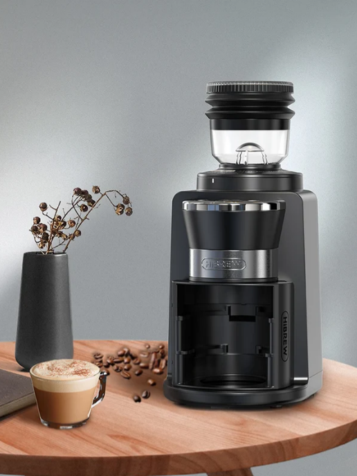 Molinillo de café negro en mesa de madera con capuchino, granos de café y florero.