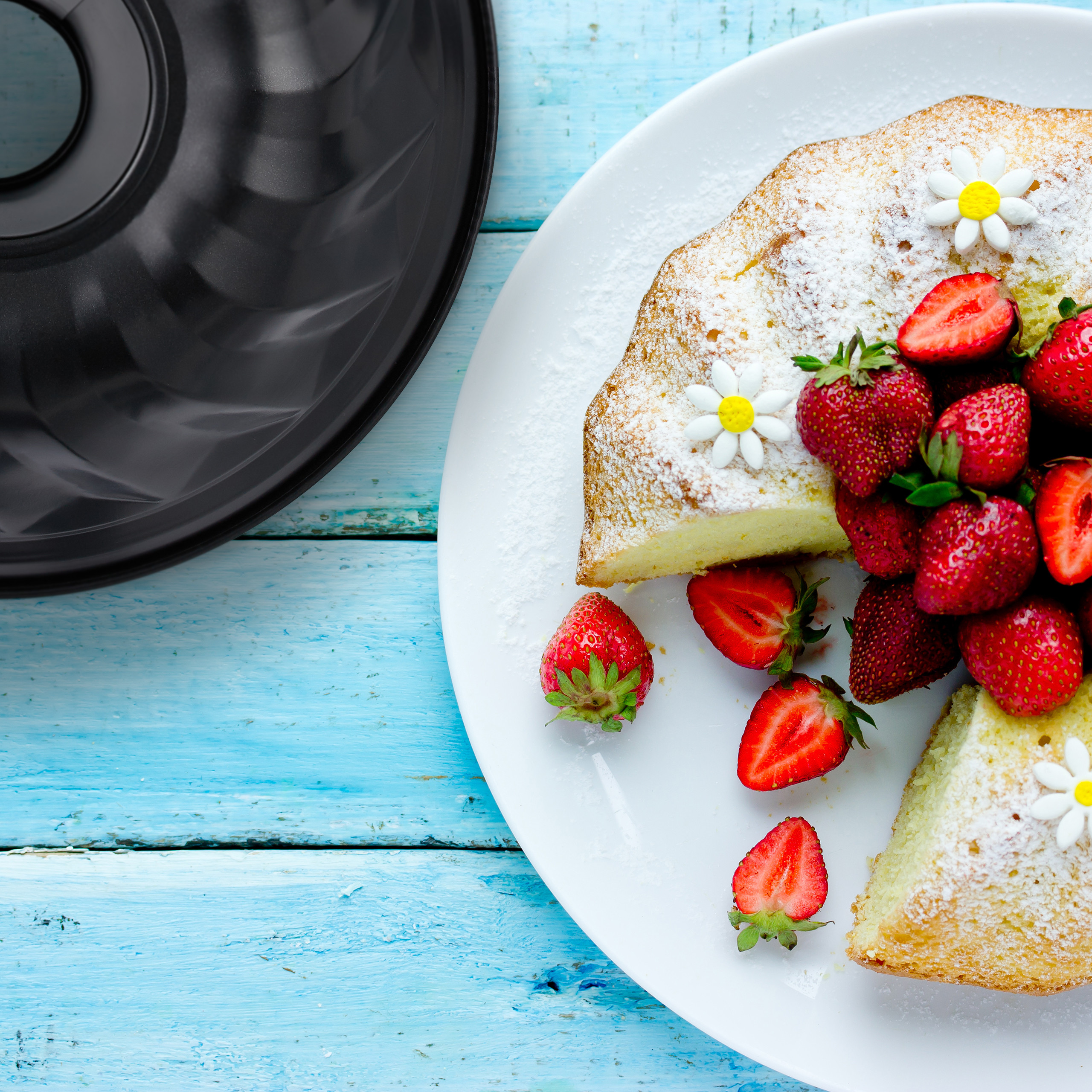 Kuchen mit Puderzucker und Erdbeeren auf weißem Teller neben Backform auf blauem Holztisch.
