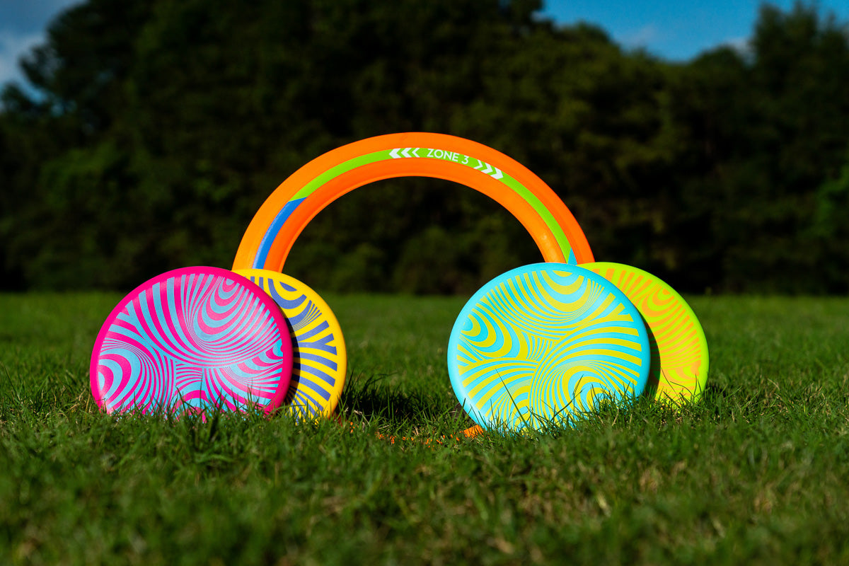 Frisbee colorati e un anello arancione si trovano nell'erba verde davanti a un bosco.