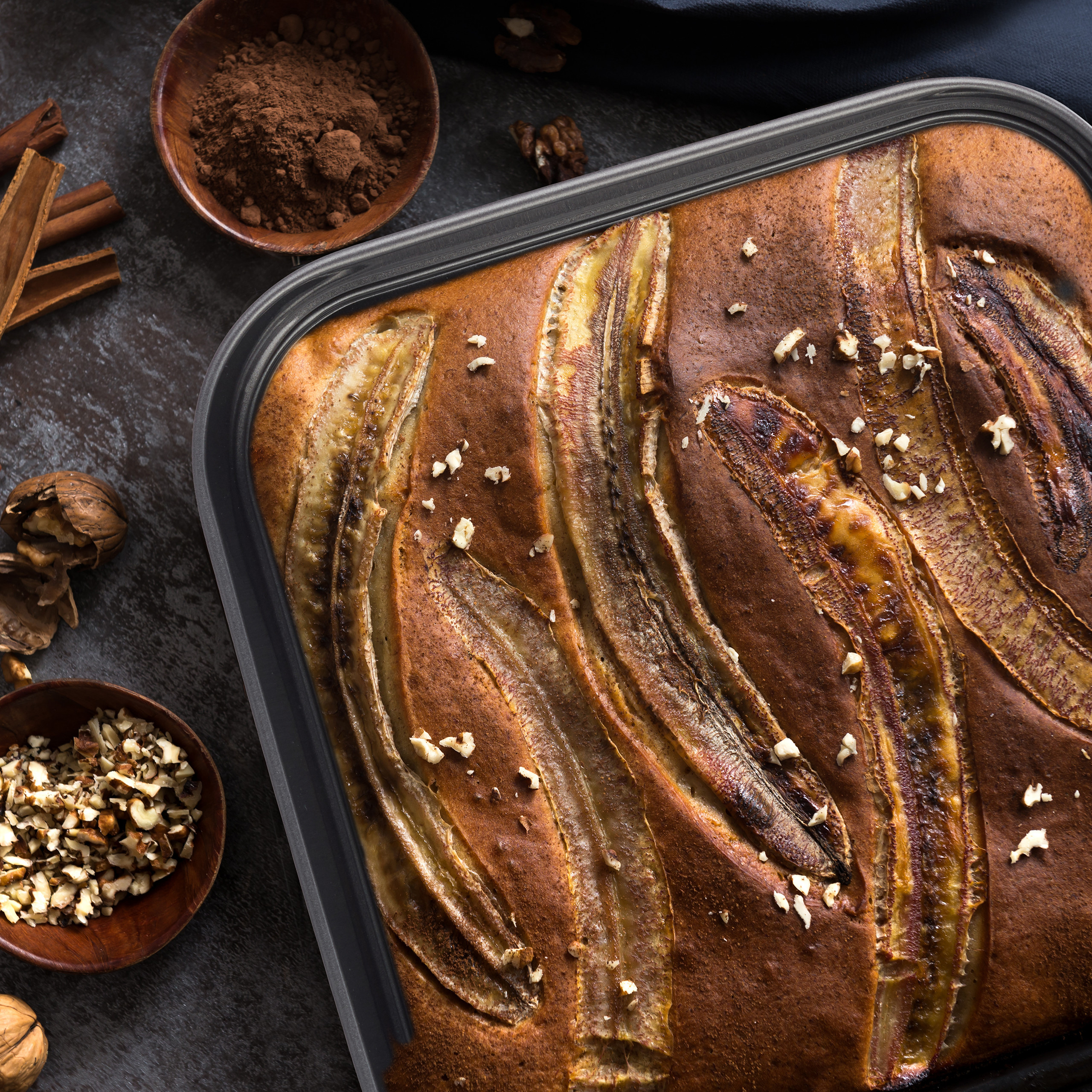 Bananenbrot in einer Backform, belegt mit Bananenscheiben und Nüssen, dazu Kakaopulver und Zimt.