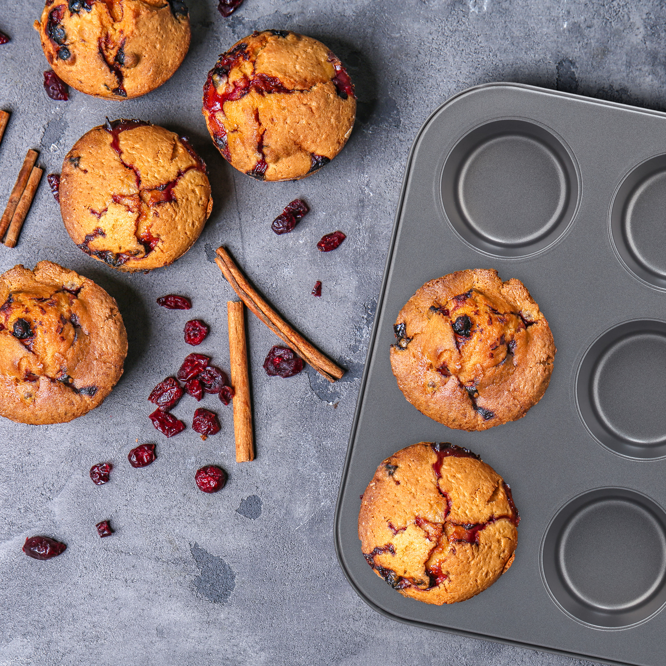 Frisch gebackene Muffins mit Cranberries und Zimtstangen neben einem grauen Muffinblech auf dunklem Untergrund.