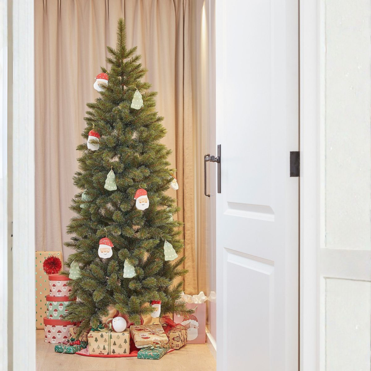 A decorated Christmas tree with ornaments and wrapped gifts beneath stands in a room viewed through an open white door.