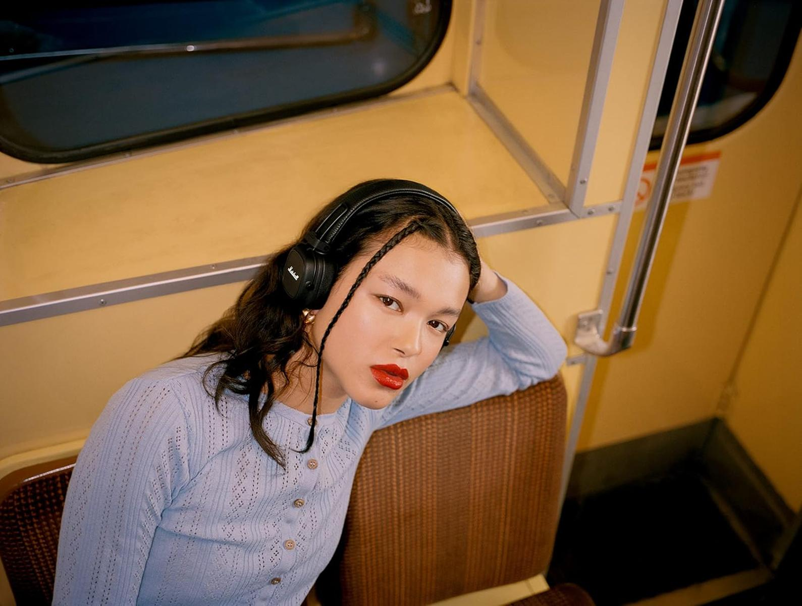 Una giovane donna con cuffie nere e maglione blu siede in metropolitana, guardando la fotocamera.
