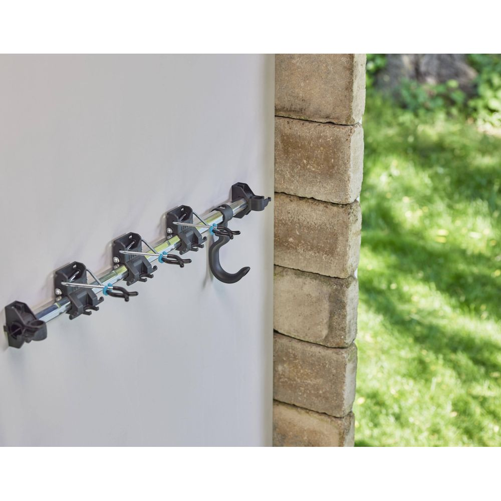 Wandmontierte Werkzeugaufhängung mit schwarzen Klemmen und Haken an einer hellen Wand neben Ziegeln und Garten.