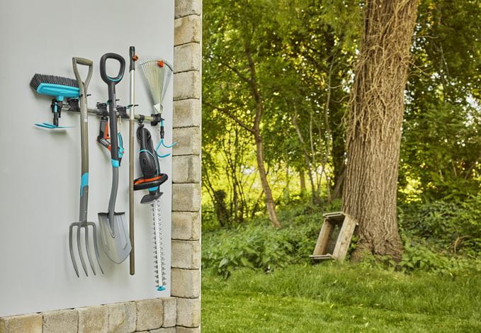 Gartengeräte hängen ordentlich an einer Wand im Freien, im Hintergrund ein grüner Garten mit Bäumen.