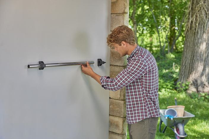 Ein blondherziger Mann im karierten Hemd montiert eine Metallstange an einer grauen Wand im Freien.
