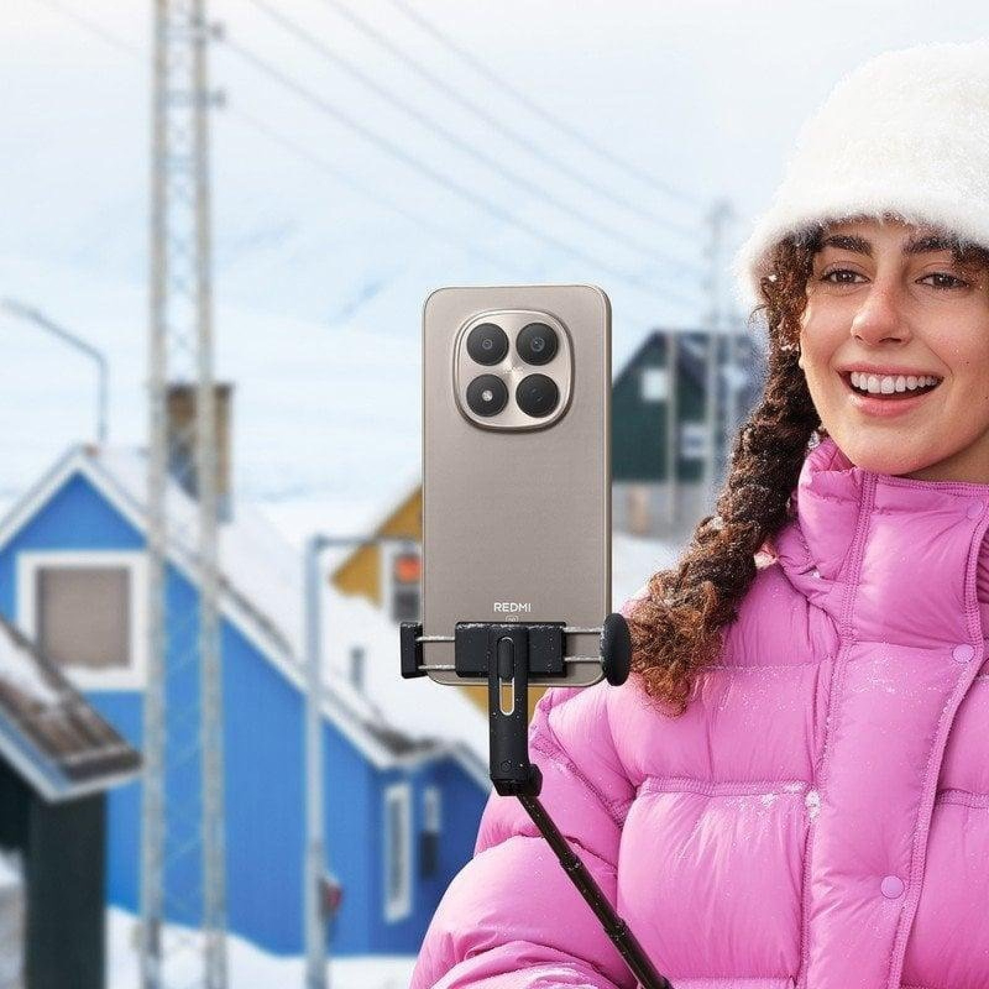 Una donna sorridente con cappotto rosa e berretto bianco tiene uno smartphone su un selfie stick con sfondo innevato.