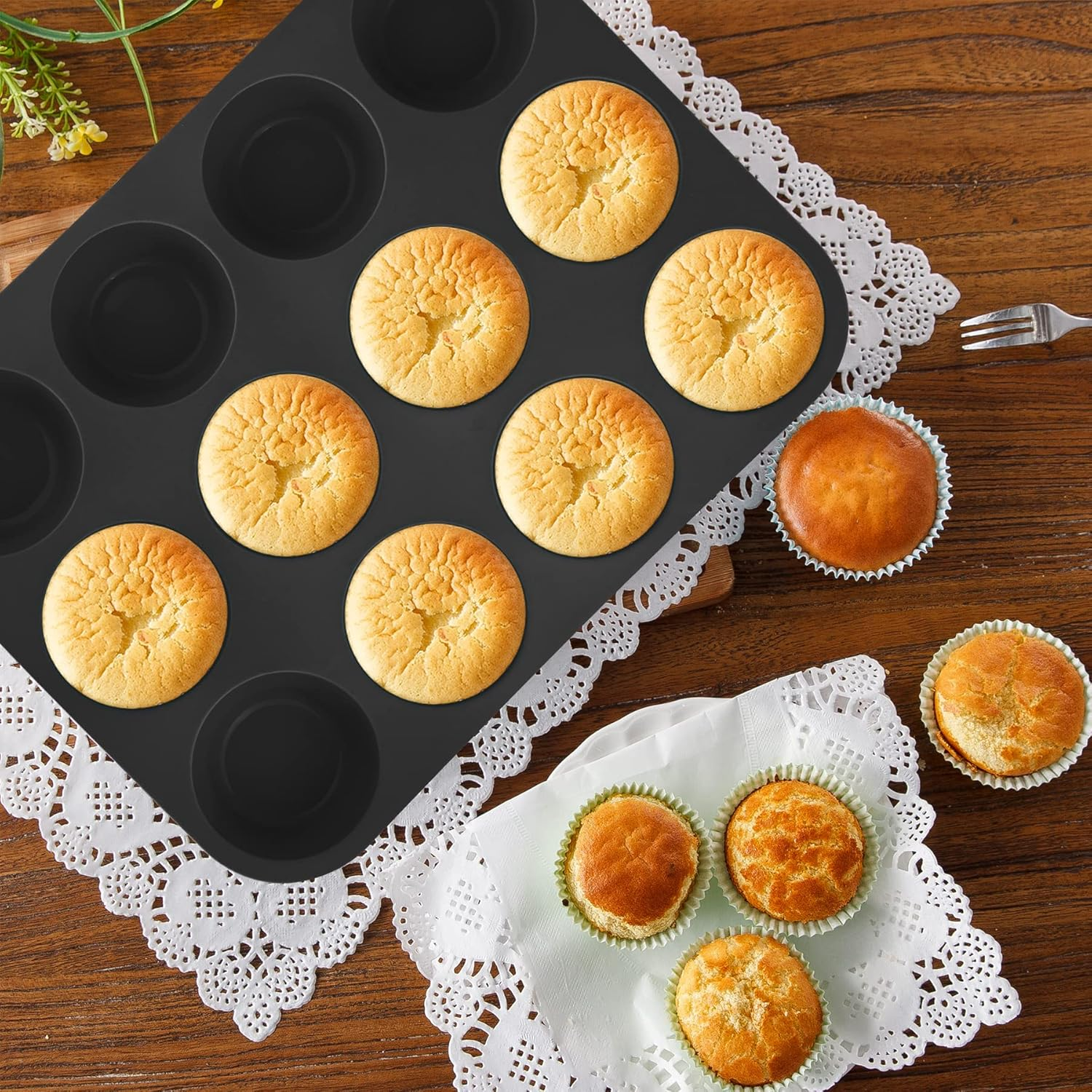 Ein schwarzes Muffinsbackblech mit gebackenen Muffins liegt auf einem Holztisch mit weißer Spitzenunterlage.