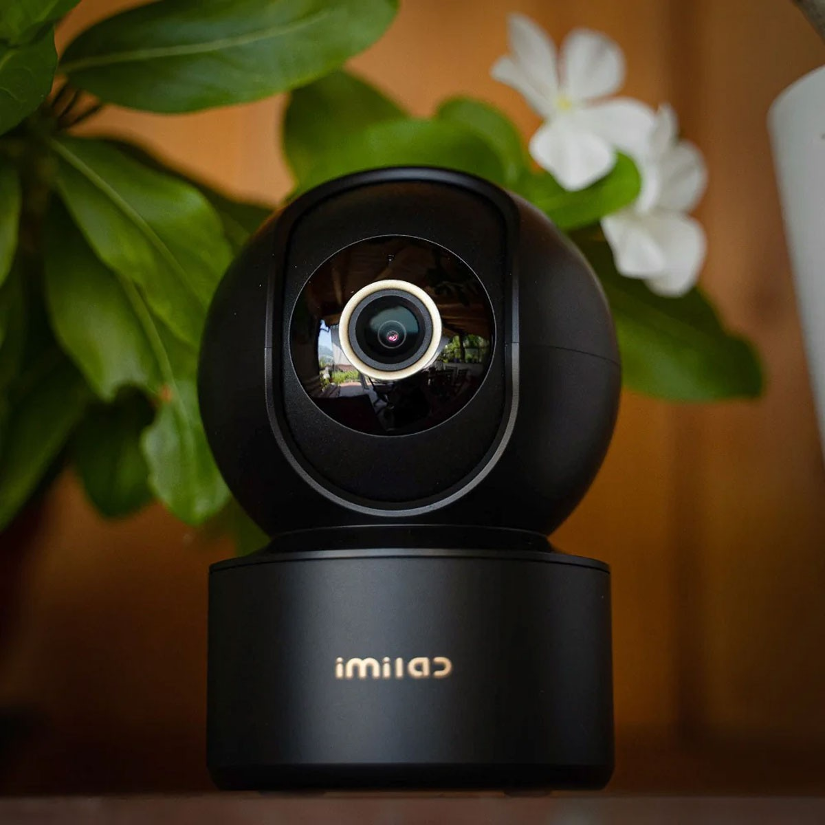 Una telecamera di sicurezza nera IMILAB poggia su una superficie davanti a piante verdi e fiori bianchi.