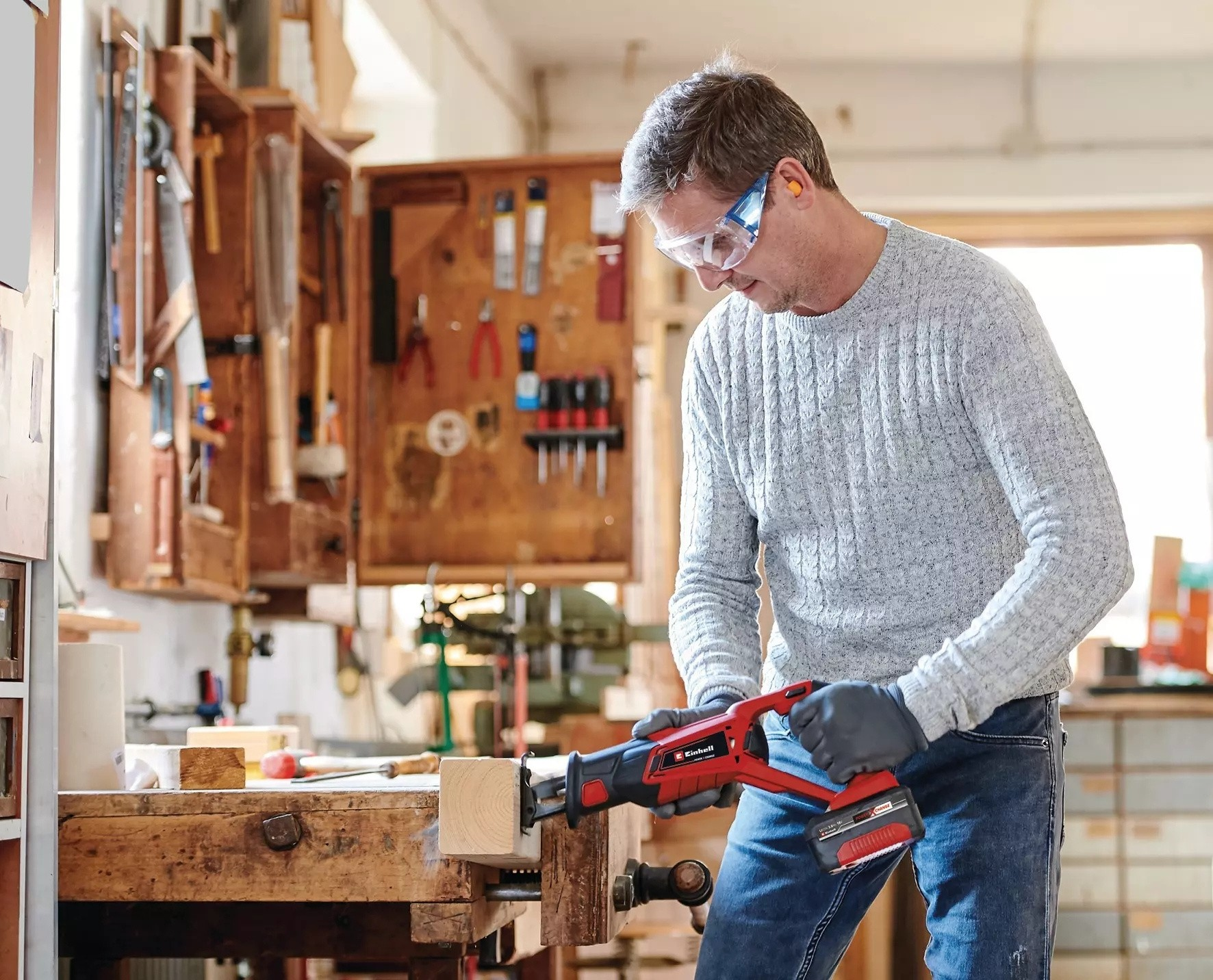 Een vakman met een veiligheidsbril zaagt een stuk hout met een elektrische zaag in een houten werkplaats.
