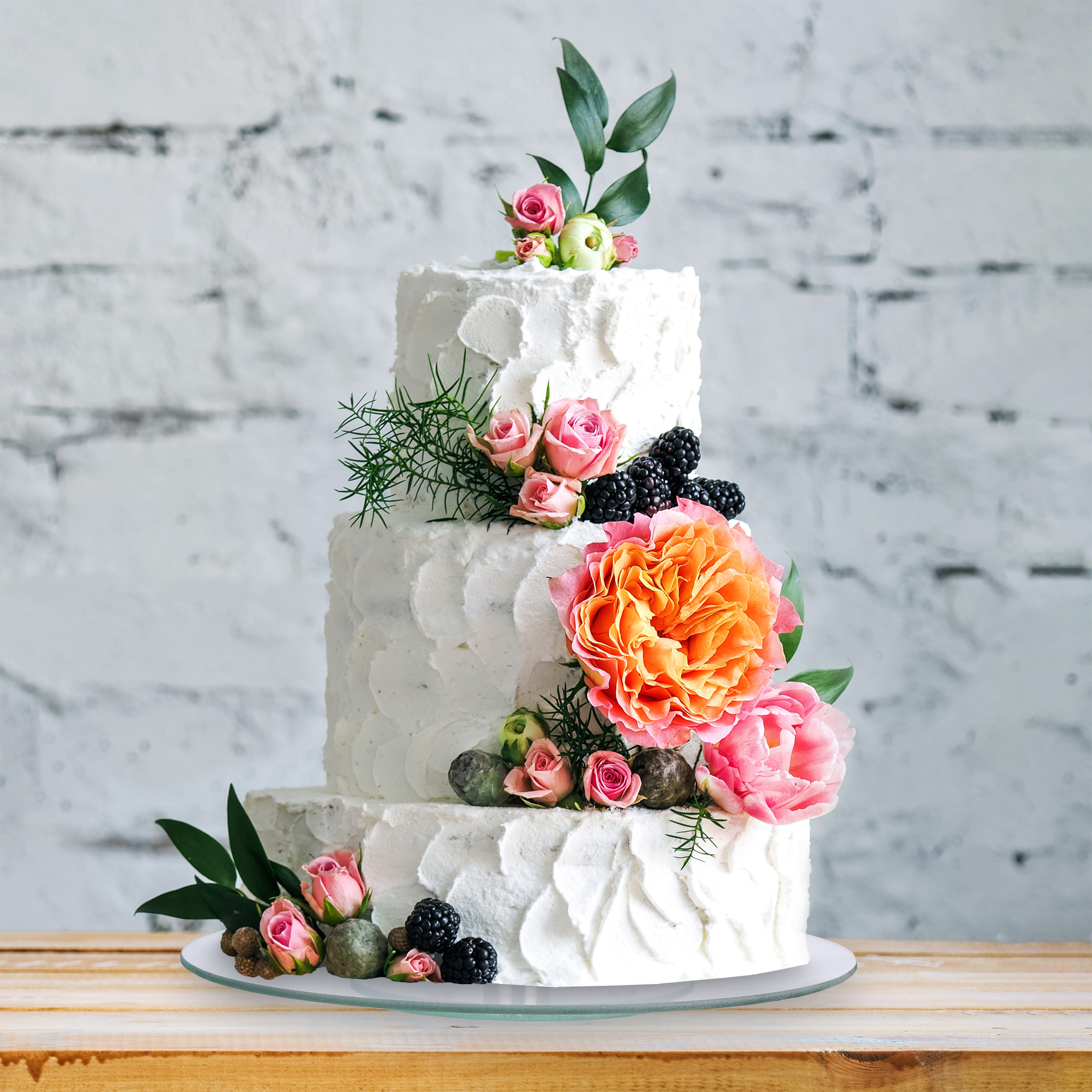 Eine dreistöckige weiße Torte, verziert mit rosa Rosen, einer großen orangefarbenen Blüte und frischen Brombeeren.