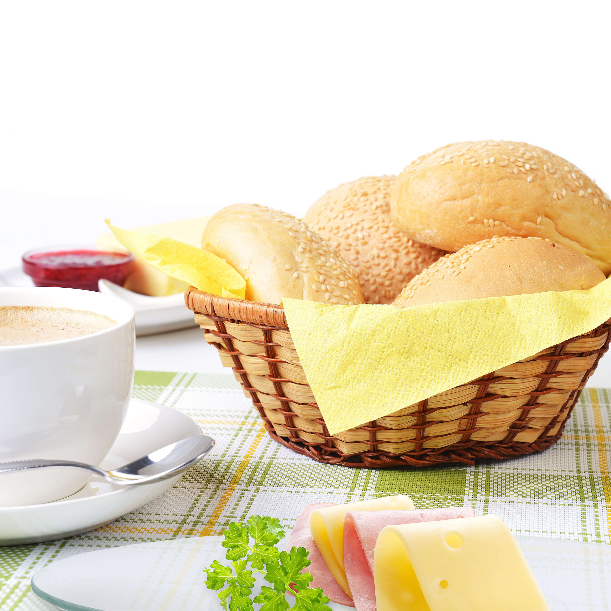 Ein Korb mit Sesambrötchen, eine Tasse Kaffee, Käse, Schinken und Marmelade auf einem karierten Platzdeckchen.