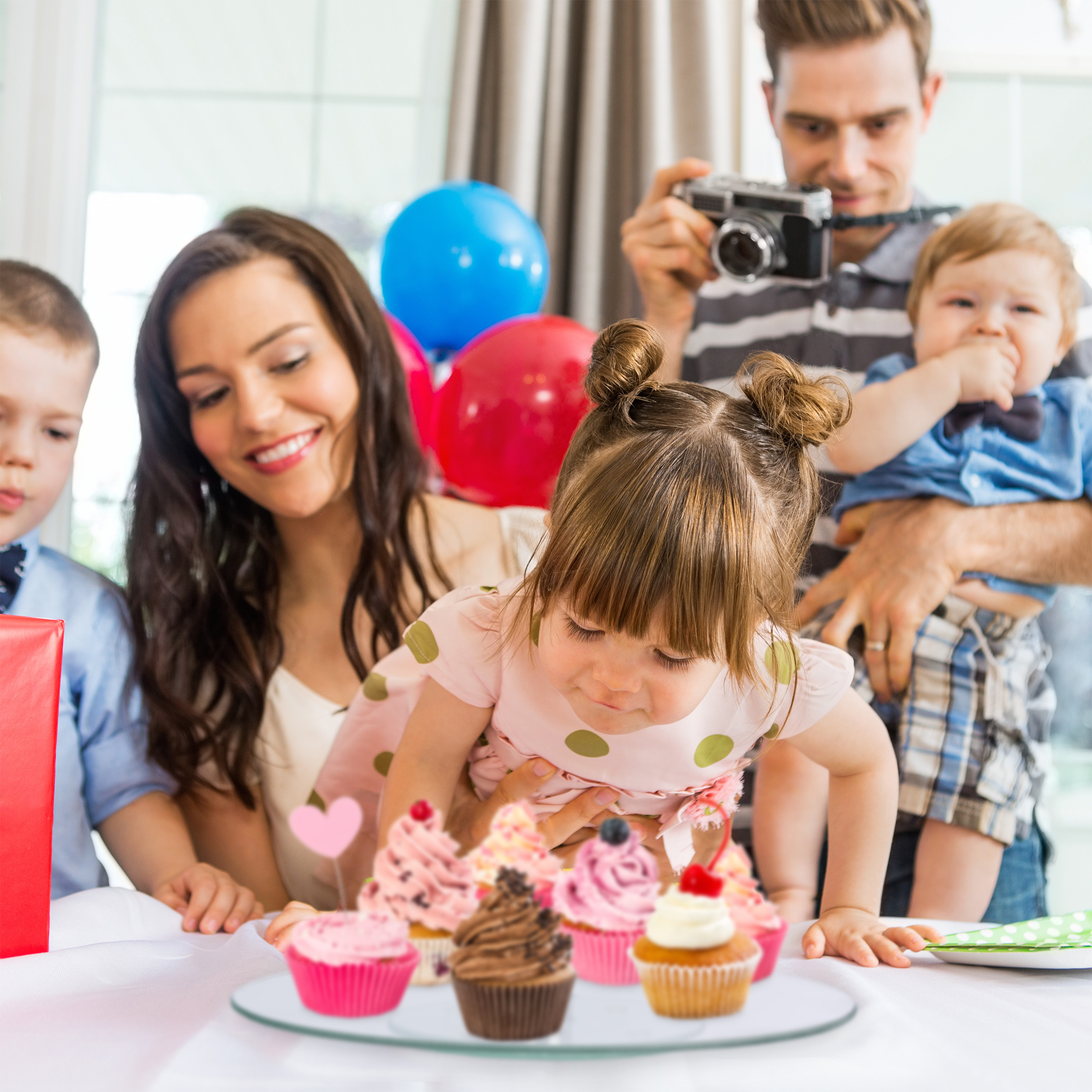 Eine Familie feiert Geburtstag, während ein Mädchen Cupcakes auf einem Tisch betrachtet und der Vater Fotos macht.
