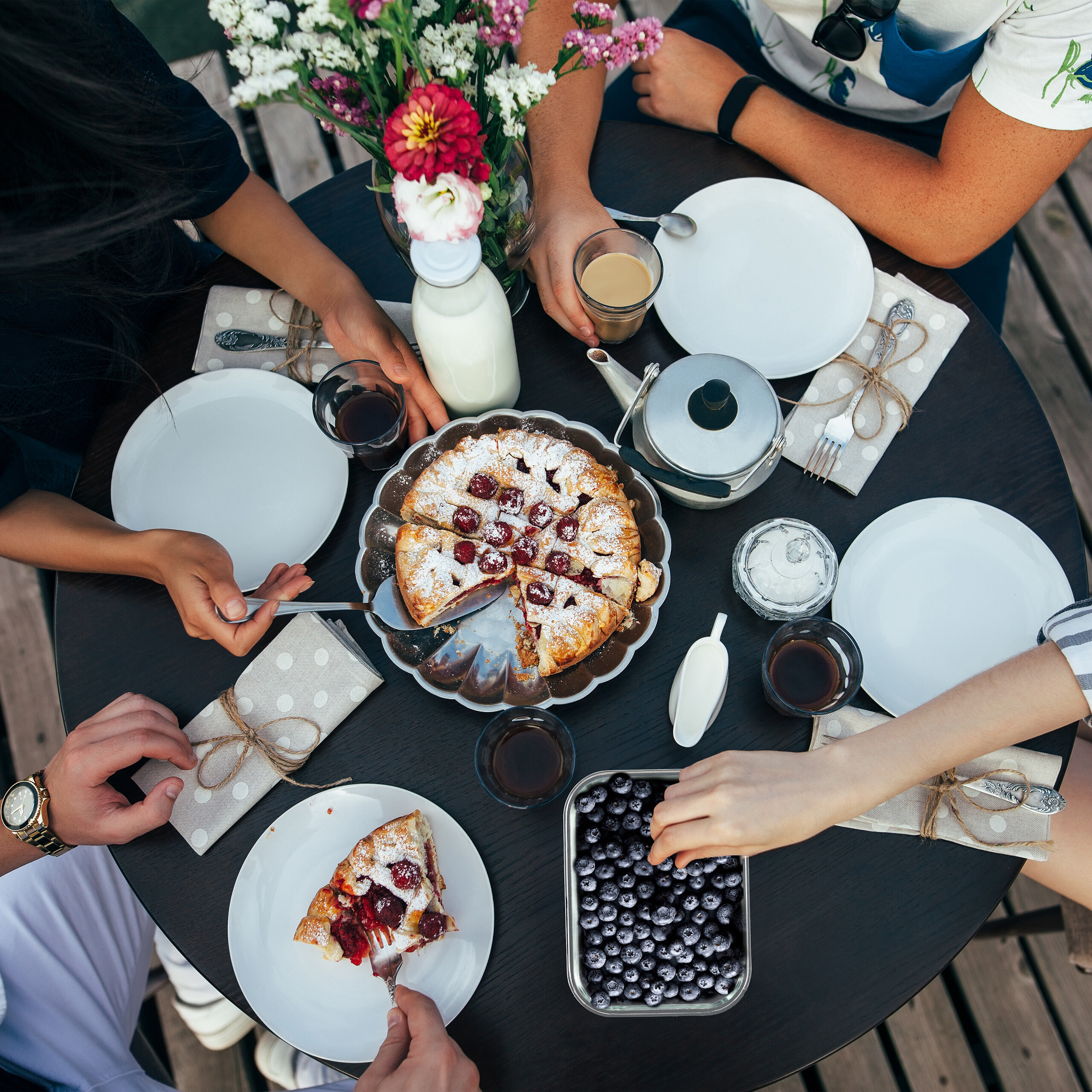 Vier Personen sitzen an einem dunklen Holztisch und essen Kirschkuchen mit Beeren, Milch und Getränken.