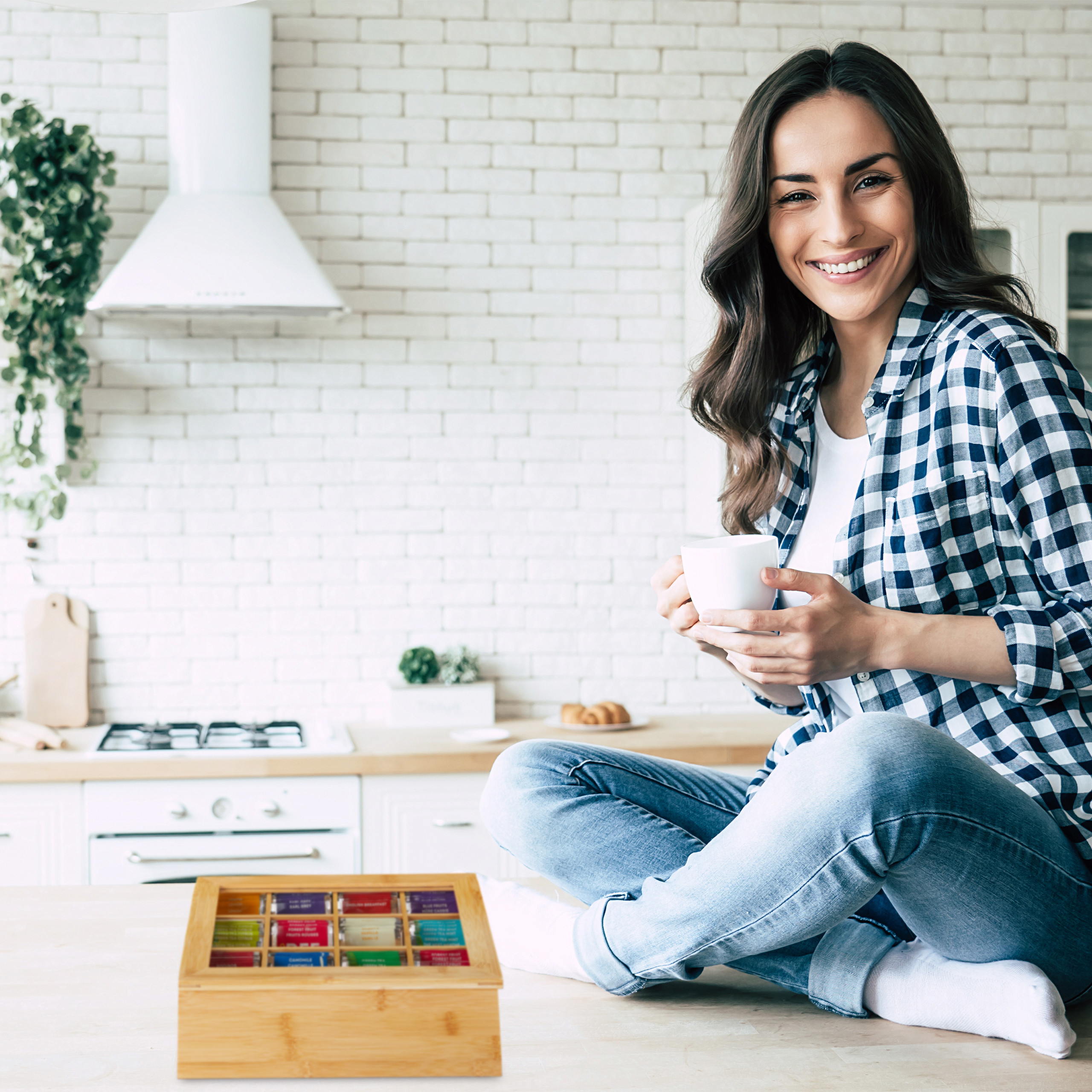 Eine lächelnde Frau in Jeans und Karohemd sitzt auf der Küchenzeile und hält eine weiße Tasse neben einer Tee-Box.