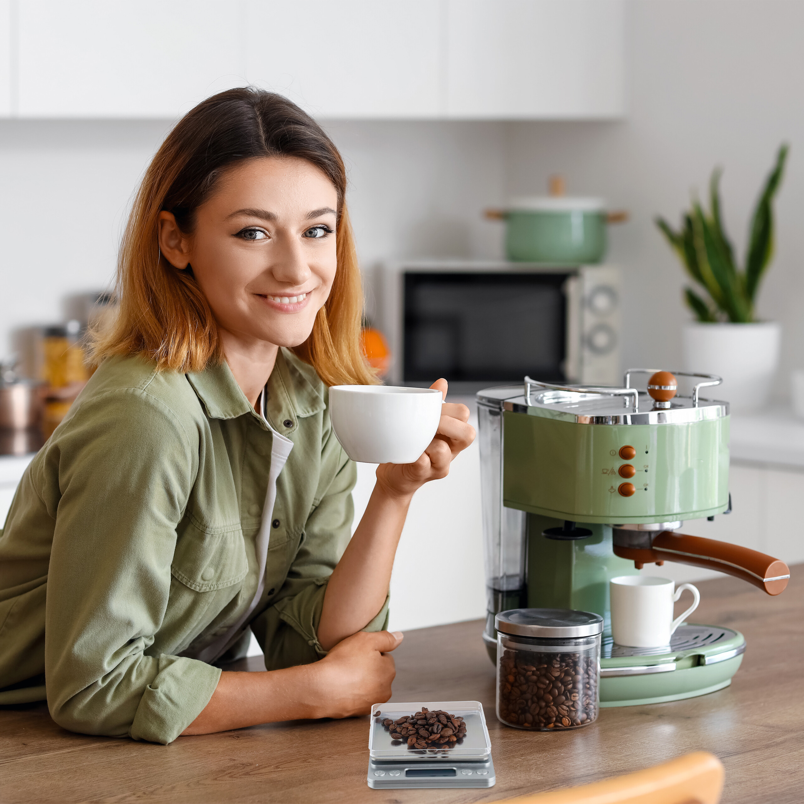 Una donna sorridente con camicia verde tiene una tazza bianca in una cucina moderna con macchina da caffè verde.
