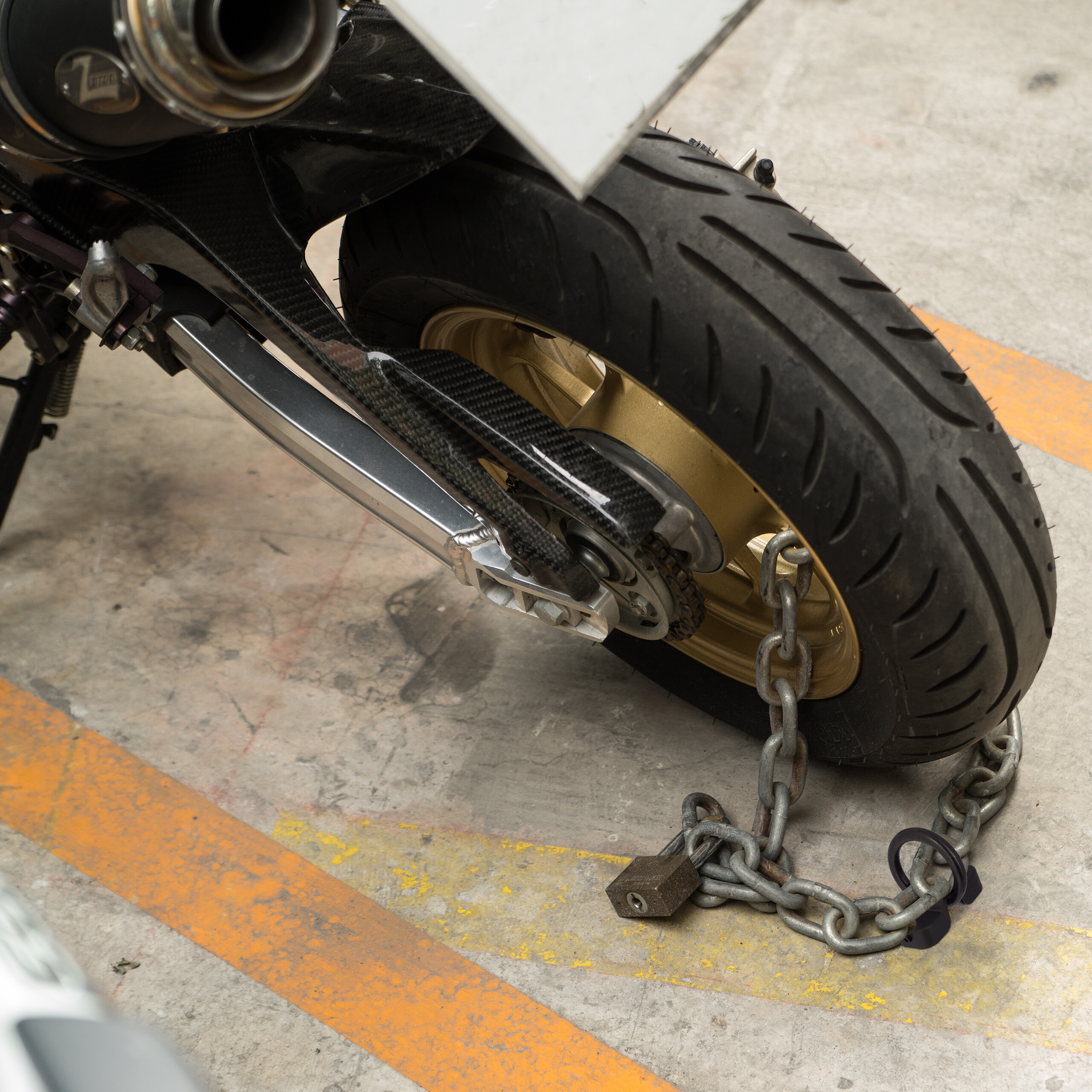 Ein Motorradhinterreifen ist mit einer schweren Kette und einem Vorhängeschloss auf einem Betonboden gesichert.