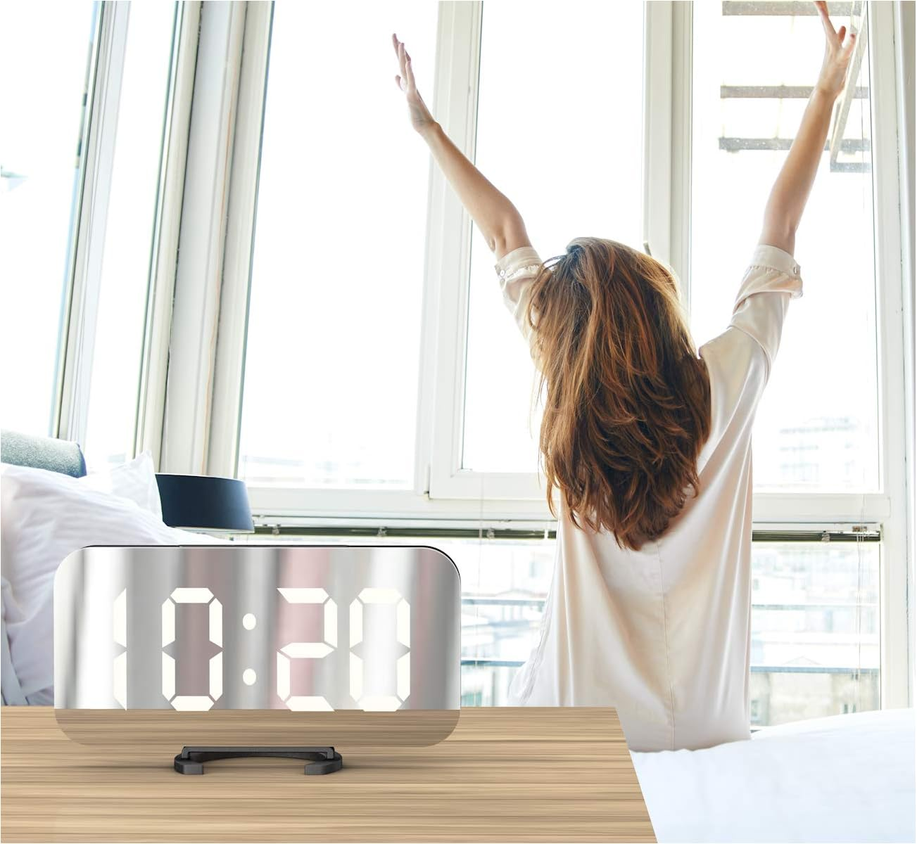 Eine Frau streckt sich morgens im Bett, im Vordergrund steht ein Digitalwecker.