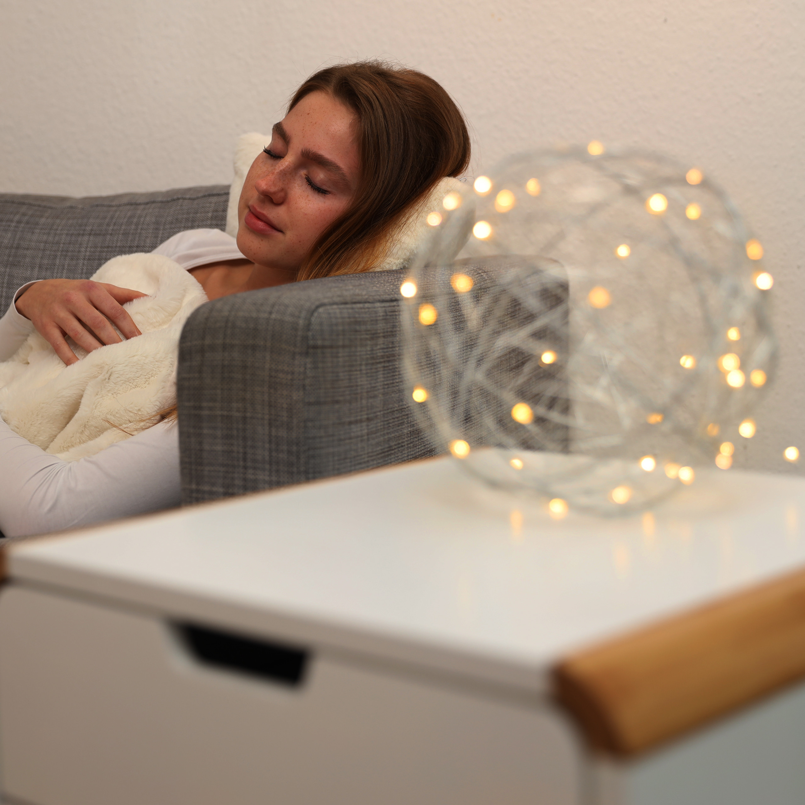 Eine junge Frau schläft friedlich auf einem grauen Sofa, zugedeckt mit einer weißen Decke, neben einer leuchtenden Lichtkugel.