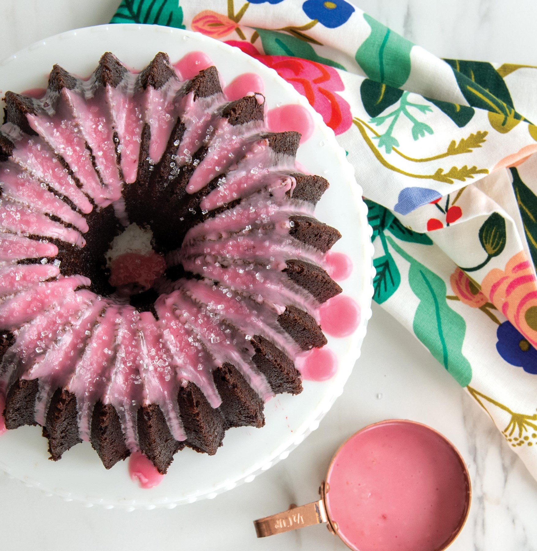 Çiçekli bez ve ölçü kabı yanında pembe soslu ve şekerli çikolatalı bundt kek.