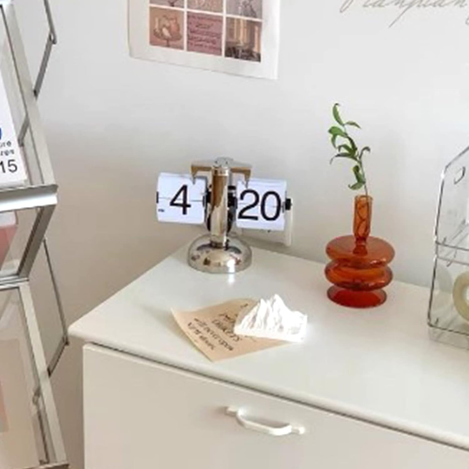 Ein weißer Tisch mit einer Flip-Uhr, die 4:20 anzeigt, einer orangen Glasvase mit Pflanze und einer Papiernotiz.