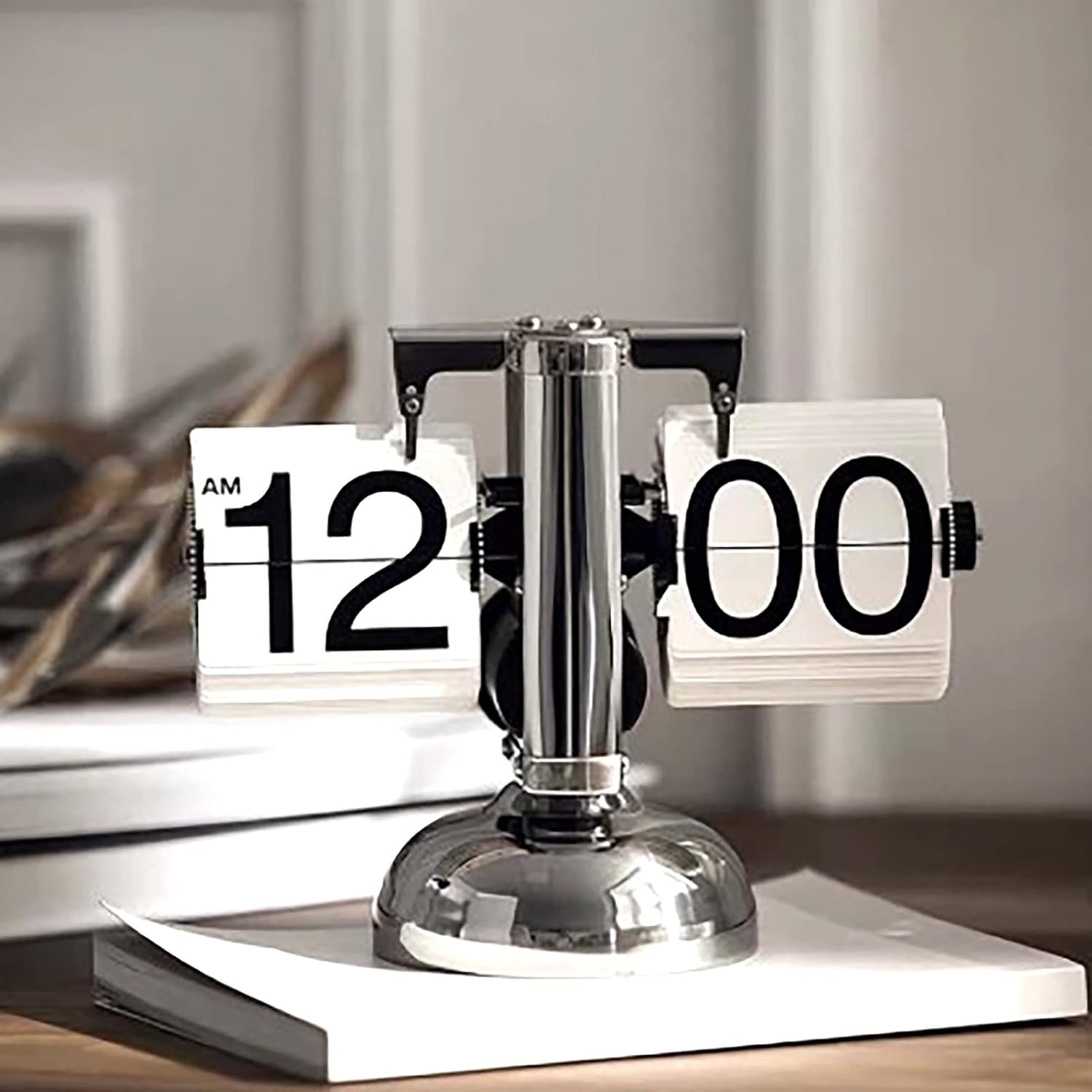 Eine silberne Retro-Flip-Uhr mit schwarzem Zifferblatt auf einem Buch, zeigt 12:00 Uhr an.