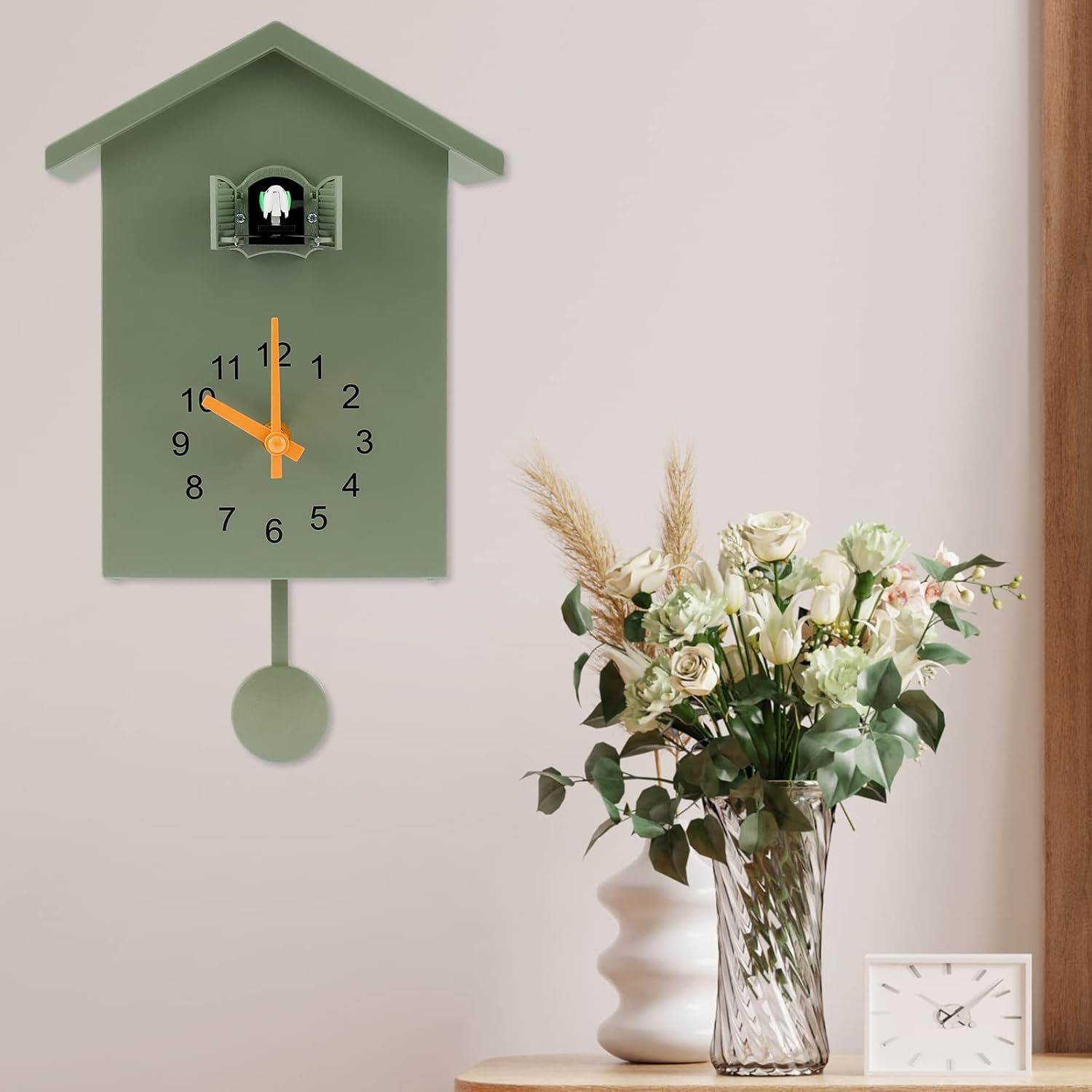 Eine grüne Kuckucksuhr hängt an der Wand neben einer Glasvase mit weißen Blumen und einer kleinen Uhr auf einem Holztisch.