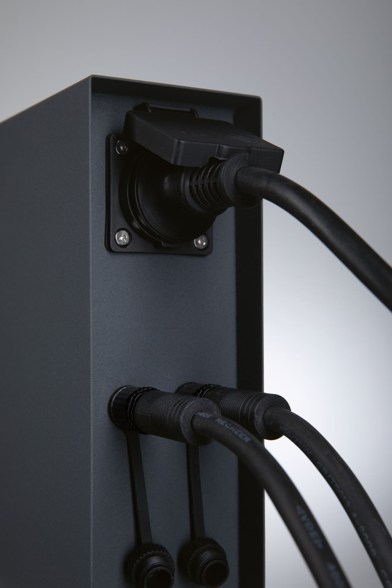 Eine schwarze Steckdosenleiste mit einem eingesteckten Kabel oben und zwei Kabeln unten vor grauem Hintergrund.