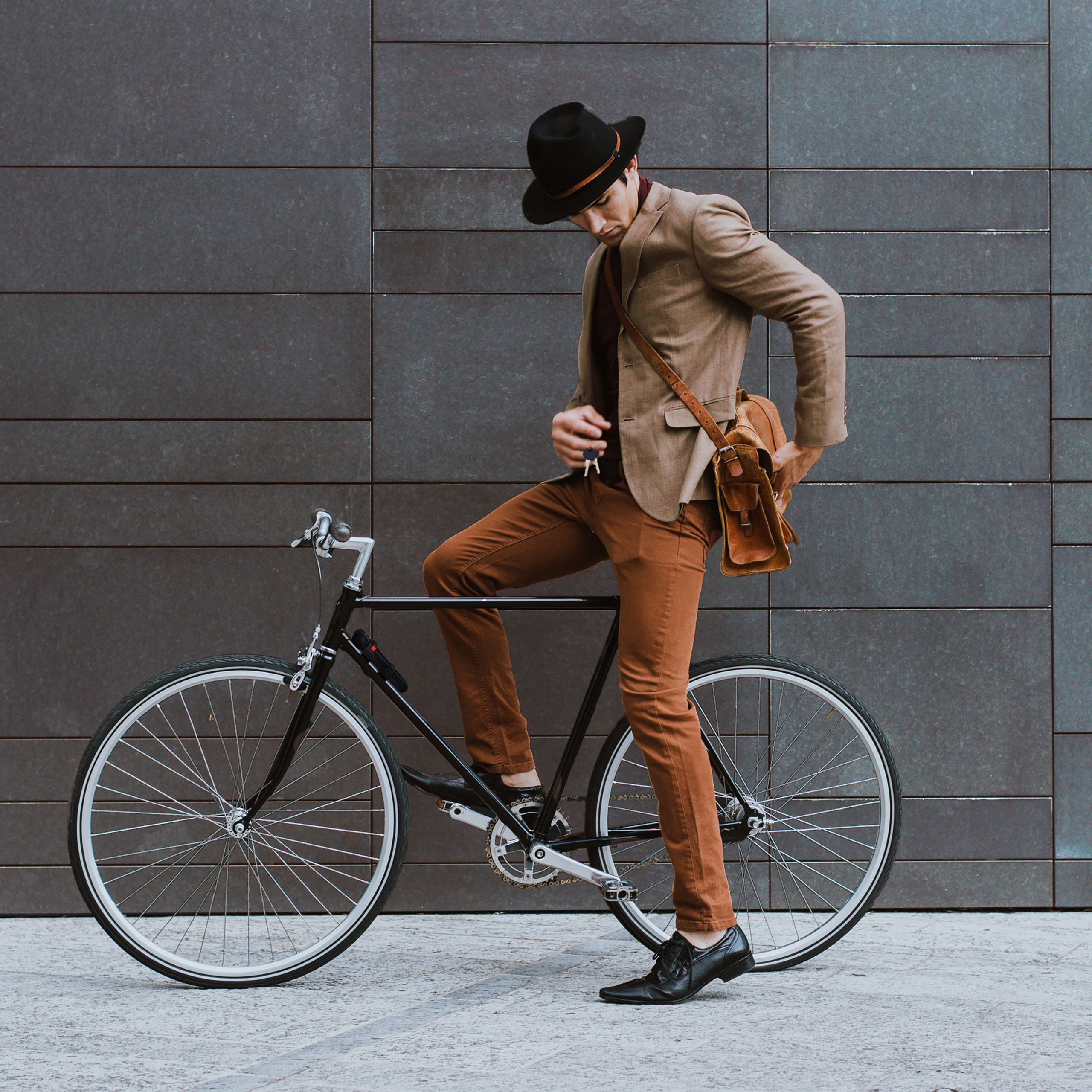 Ein stilvoller Mann im Hut mit einer Tasche und einem schwarzen Fahrrad vor einer grauen Wand.