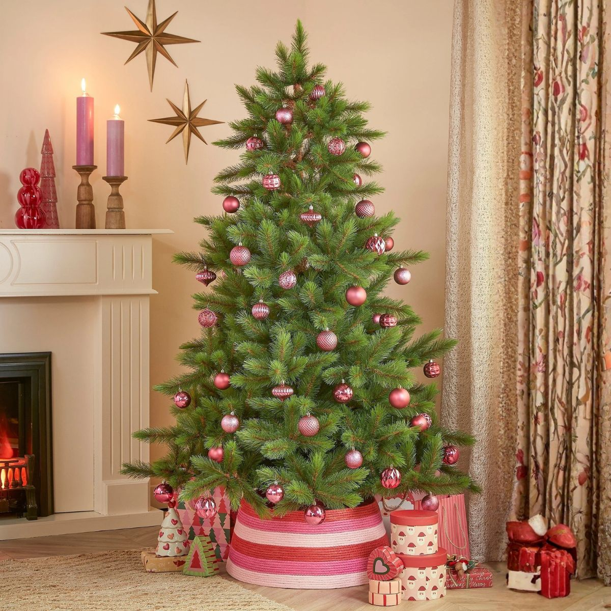 Un sapin de Noël décoré de boules roses dans un salon avec une cheminée, des bougies et des cadeaux.