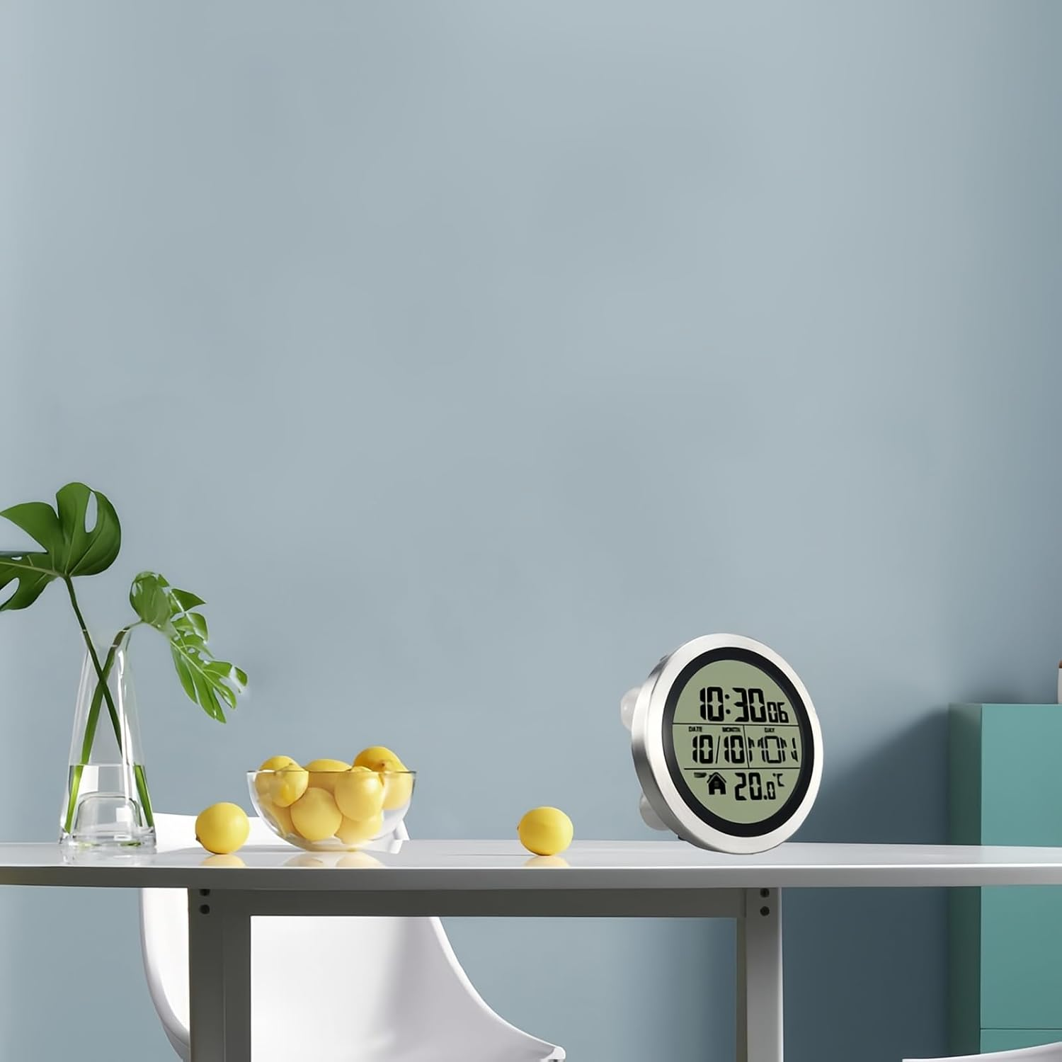 Ein weißer Tisch mit Digitaluhr, Zitronenschale und Vase mit grünen Blättern vor hellblauem Hintergrund.
