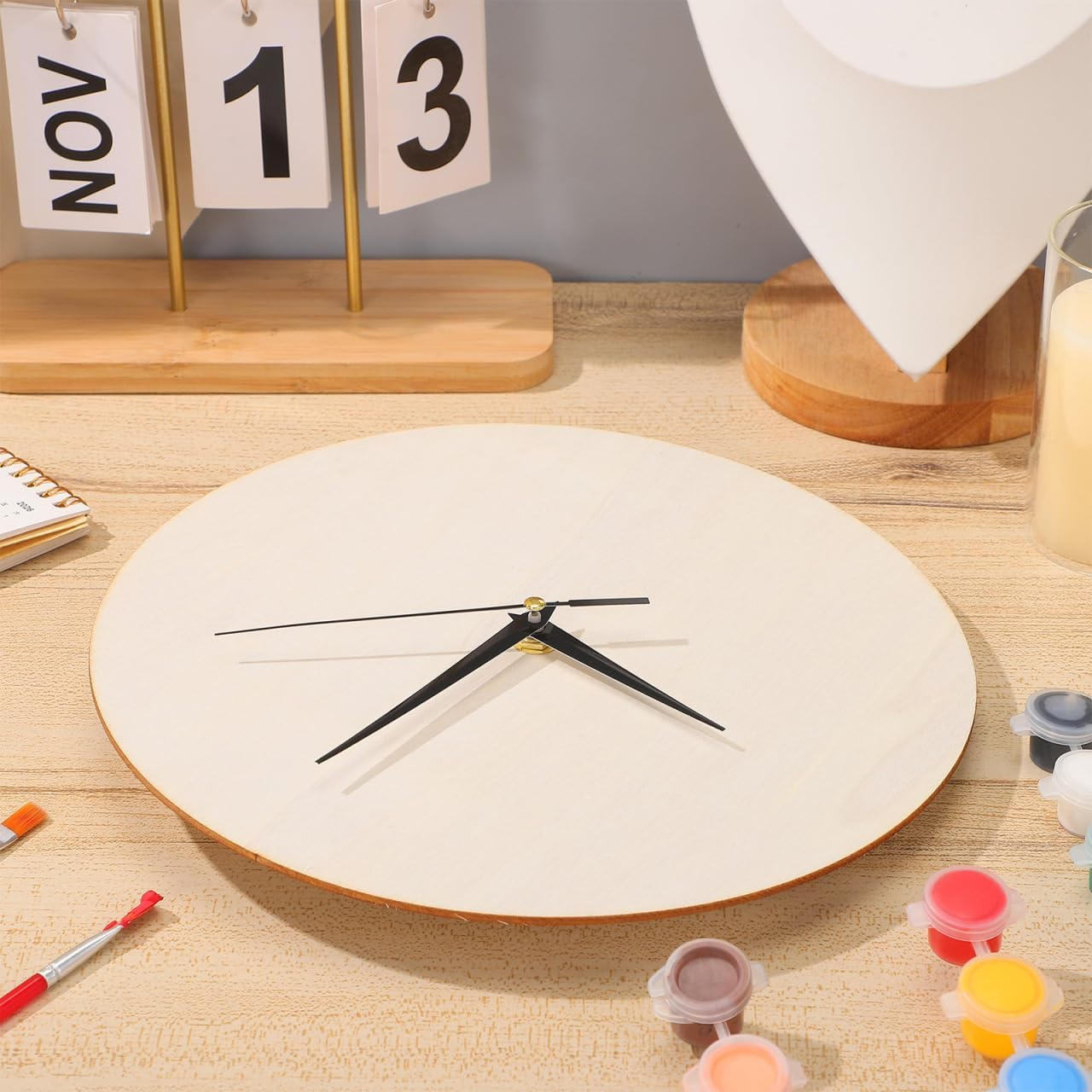 Eine hölzerne Uhr mit schwarzen Zeigern auf einem Tisch mit Farbe und einem Kalender im Hintergrund.