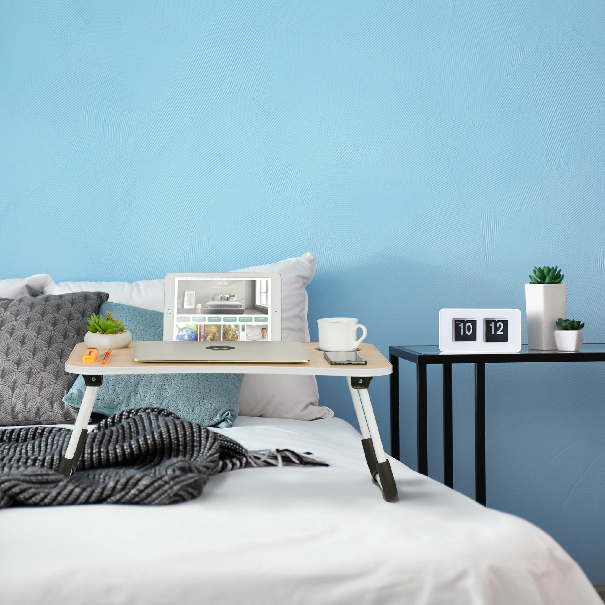 Laptoptisch auf einem weißen Bett mit Kissen, einem Tablet, Tasse und kleinen Pflanzen.