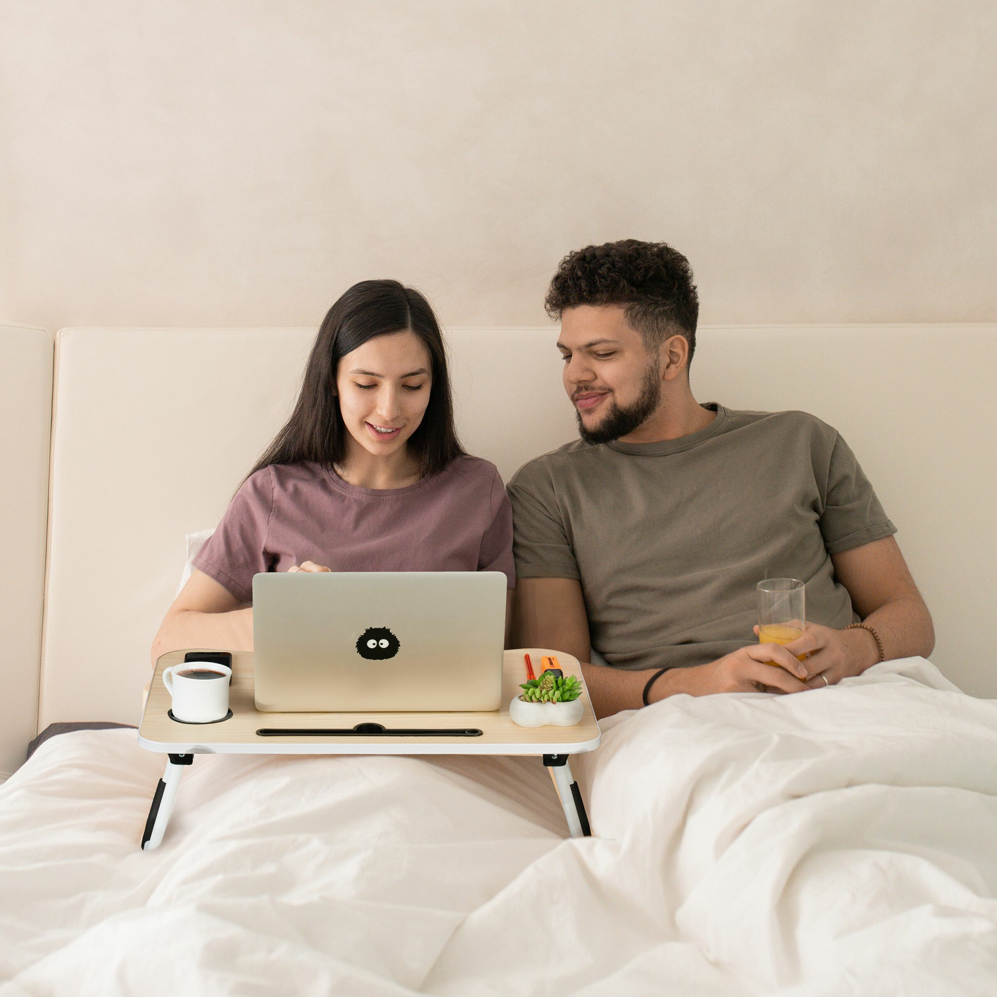 Paar sitzt im Bett mit Laptop auf einem Betttisch, sie tippt, er hält ein Glas Orangensaft.