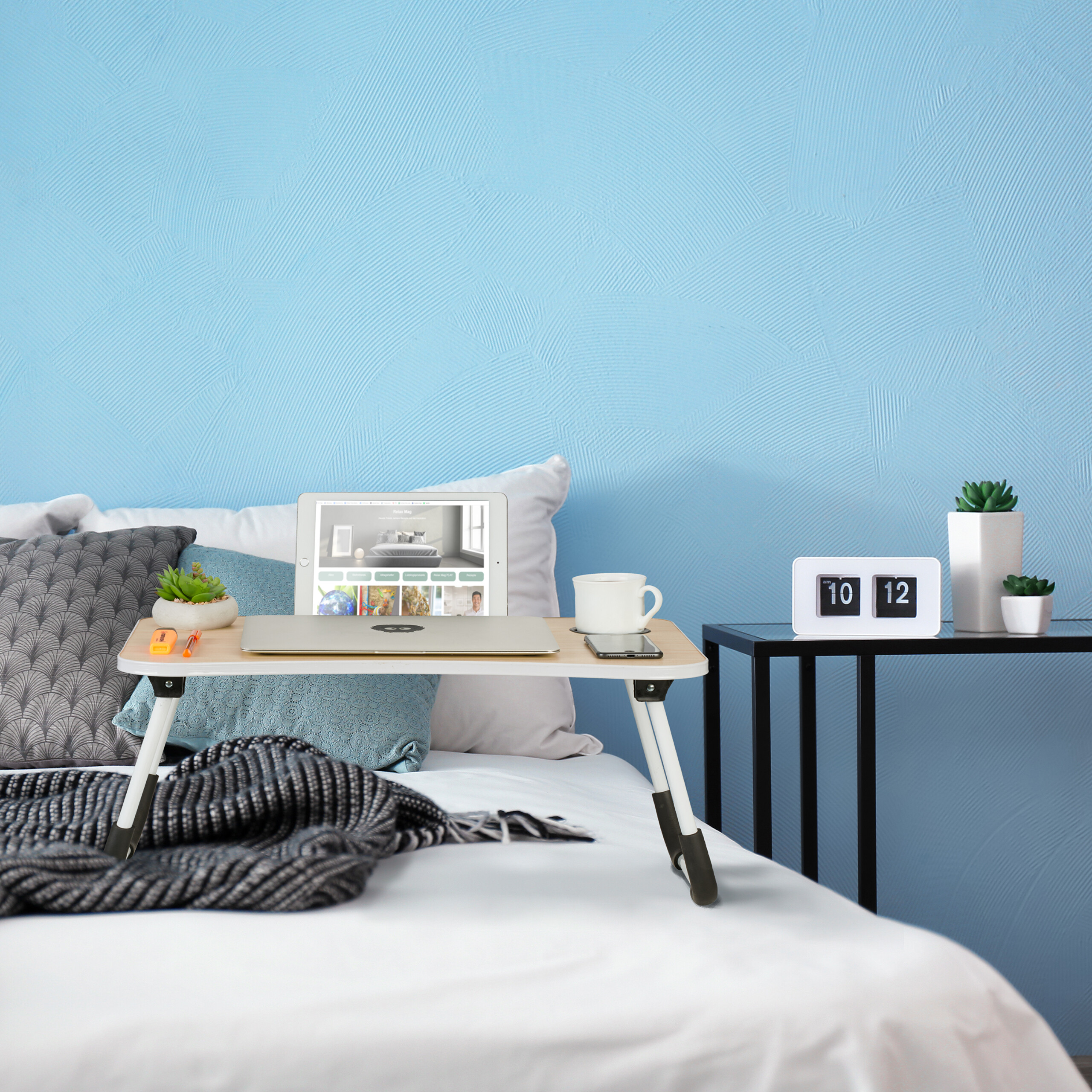 Ein Bett mit einem Laptop auf einem kleinen Tisch, Kissen, einer Tasse und einer Wand im Hintergrund.
